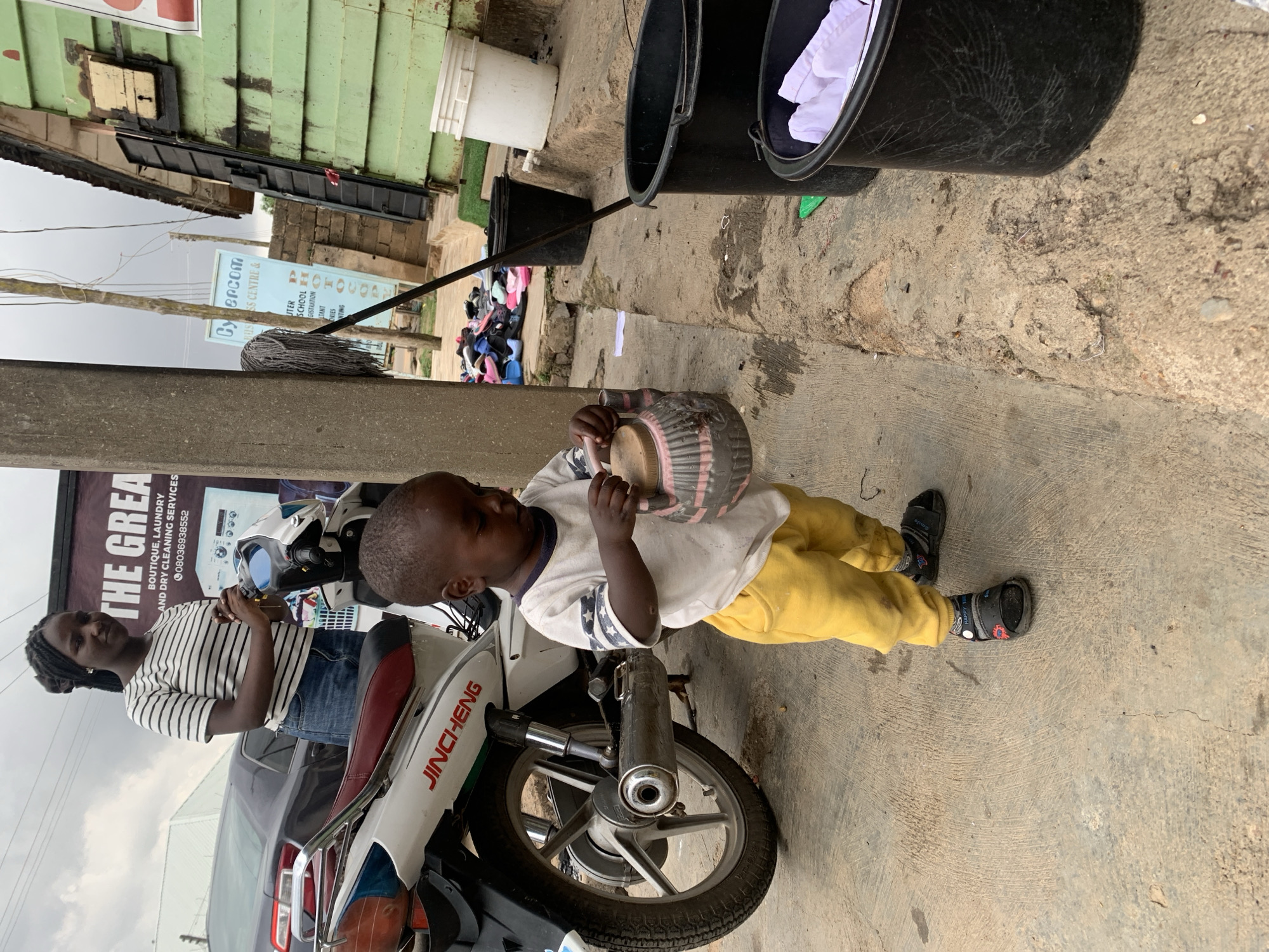 The image depicts an outdoor scene in Jos, Nigeria, likely a street in a commercial area. In the foreground, a young child, appearing to be a toddler, stands on a concrete ground wearing yellow pants and sandals. The child is holding a beige and pink patterned container. To the left of the child is the rear portion of a white motorcycle with red accents, bearing the "JINCHENG" logo. Behind the motorcycle, a young woman with braided hair, wearing a striped t-shirt and denim shorts, stands with her hands clasped. She is looking slightly to her right. A large banner with "THE GREAT" and other text is visible behind them. In the background, there is a green wooden building with a weathered facade, a bucket, and several other large black plastic containers, one of which holds white cloth items. Scattered footwear can be seen near the building. A utility pole with wires runs vertically through the scene. The overall environment suggests a bustling, somewhat disorganized urban setting.