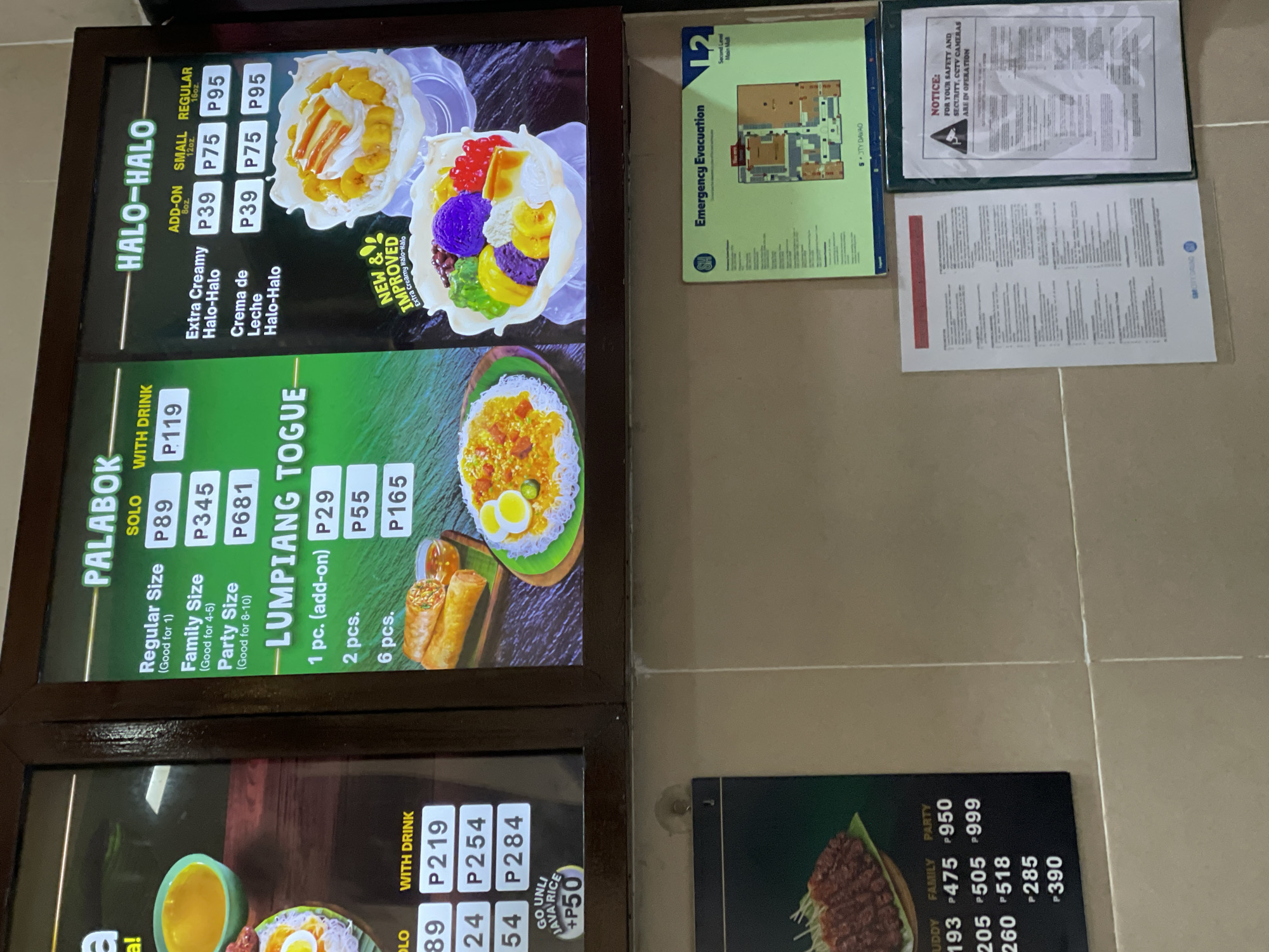 This image captures a section of a commercial establishment, likely a fast-food counter or restaurant, located within the SM City Davao mall on its Second Level (L2 Main Mall) in Davao City, Philippines, as indicated by the emergency evacuation map.
The upper portion of the scene is dominated by three vertically aligned menu boards. The left board, partially cropped, displays prices in Philippine Pesos (P), including options for "SOLO," "WITH DRINK," and "GO UNLI JAVA RICE +P50." The central menu board advertises "PALABOK" with prices for Regular Size (P89 solo, P119 with drink), Family Size (P345 solo), and Party Size (P681 solo), along with an image of the dish featuring noodles, sauce, and an egg. Below this, "LUMPIANG TOGUE" is listed at P29 for 1 pc. (add-on), P55 for 2 pcs., and P165 for 6 pcs., accompanied by an image of spring rolls with dipping sauce. The rightmost menu board features "HALO-HALO," listing "Extra Creamy Halo-Halo" and "Crema de Leche Halo-Halo" each at P39 (add-on), P75 (small 8oz), and P95 (regular 16oz). Two images of Halo-Halo are shown; one with a creamy, banana-topped appearance, and another labeled "NEW & IMPROVED Extra Creamy Halo-Halo" displaying a colorful mix of ingredients including purple yam, red beans, and various jellies in a clear bowl.
The lower portion of the image shows a light-colored, possibly tiled, wall. Affixed to this wall is a rectangular "Emergency Evacuation" map displaying the SM logo and a floor plan, explicitly stating "L2 Second Level Main Mall" and "SM CITY DAVAO." Below this map, two framed notices are displayed. The notice on the right, housed in a green frame, clearly states: "NOTICE: FOR YOUR SAFETY AND SECURITY, CCTV CAMERAS ARE IN OPERATION," followed by additional text. The notice on the left, encased in a clear sleeve, also contains text and "SM CITY DAVAO" at its base. No individuals are visible within the depicted area.