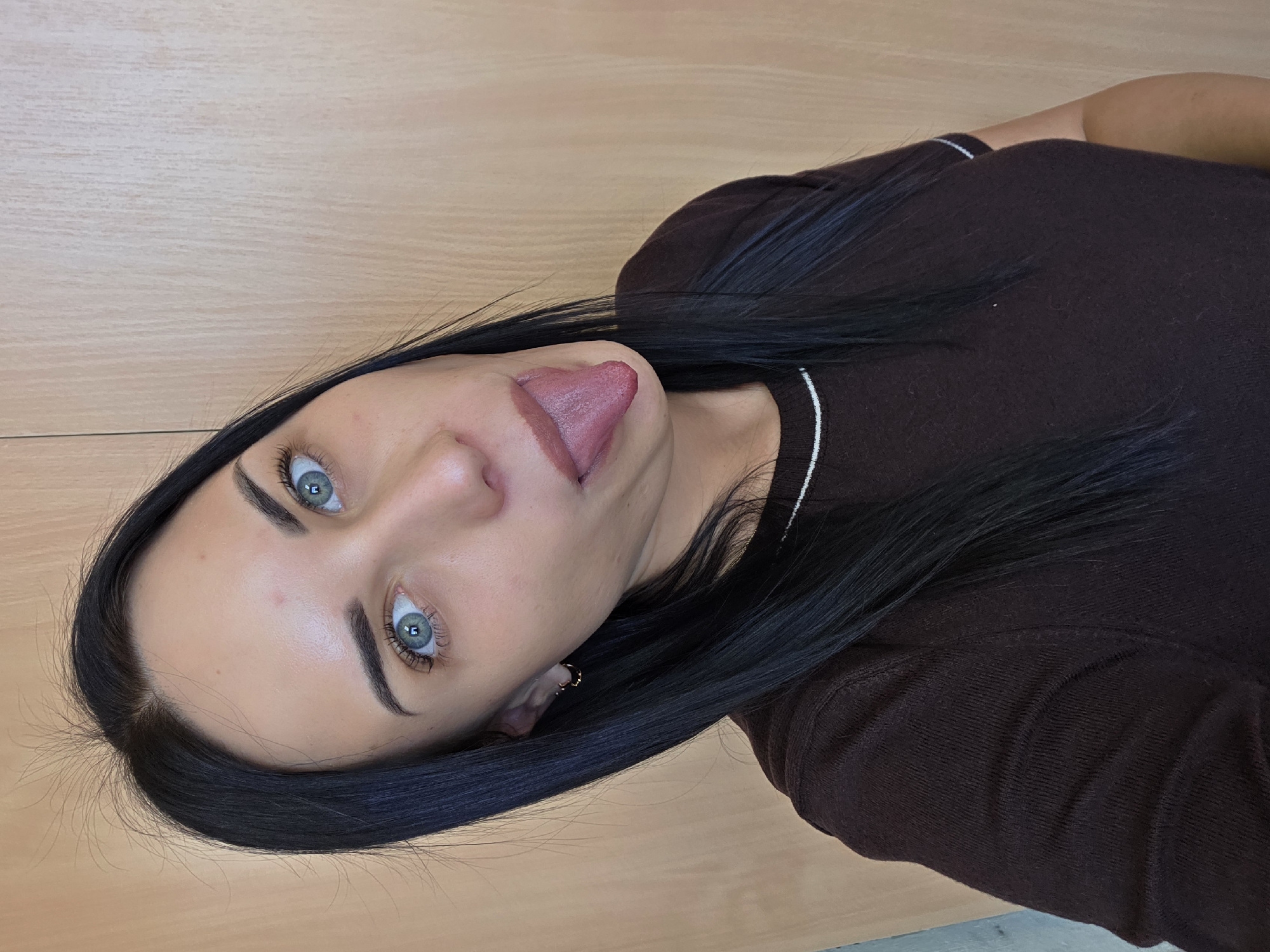 A woman with dark hair and light blue eyes is seen from a close-up, tilted angle. She is sticking her tongue out. She is wearing a brown top. The background is a light brown surface. There is no visible indication of the location being Chaikovsky, Russia.