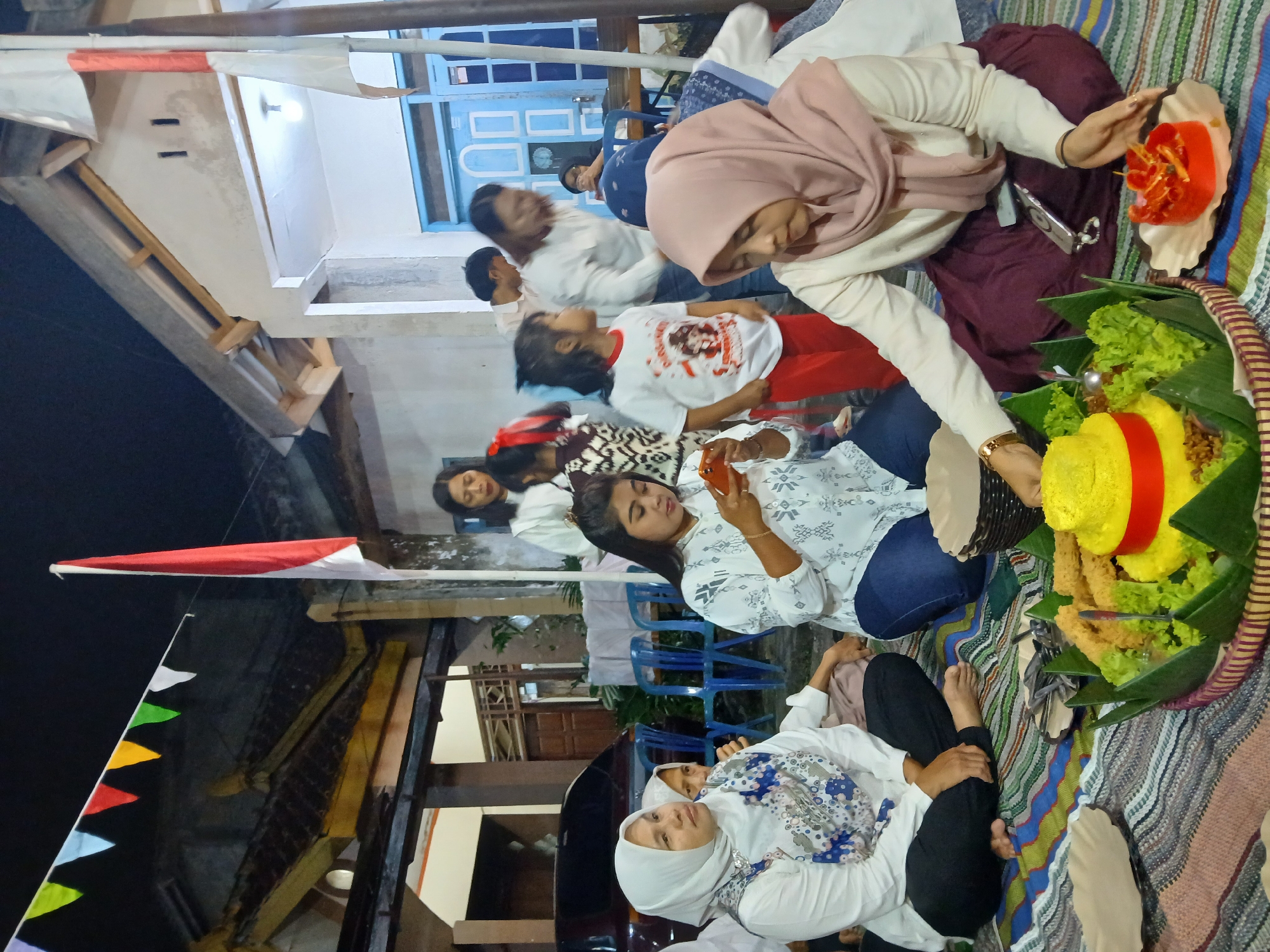This image depicts an outdoor gathering in what appears to be a residential area in Doplang, Indonesia. The scene is set at night, illuminated by artificial lights. Several people, predominantly women and children, are seated on a colorful woven mat laid out on the ground. They are dressed in casual attire, with some women wearing headscarves.

In the foreground, a traditional Indonesian cone-shaped rice cake, known as "Nasi Tumpeng," is prominently displayed on a woven tray garnished with banana leaves and lettuce. Accompanying the rice cake are various side dishes and condiments, including fried snacks and a small red dish with skewers.

A few individuals are engaged with their mobile phones, while others are interacting with each other. In the background, a house with a visible blue door and window frames is visible, suggesting a domestic setting. A national flag of Indonesia, red and white, is raised on a pole, and decorative triangular flags are strung across the scene, indicating a celebratory or national event. A dark-colored car is parked to the left of the frame. The overall atmosphere suggests a community or family gathering.