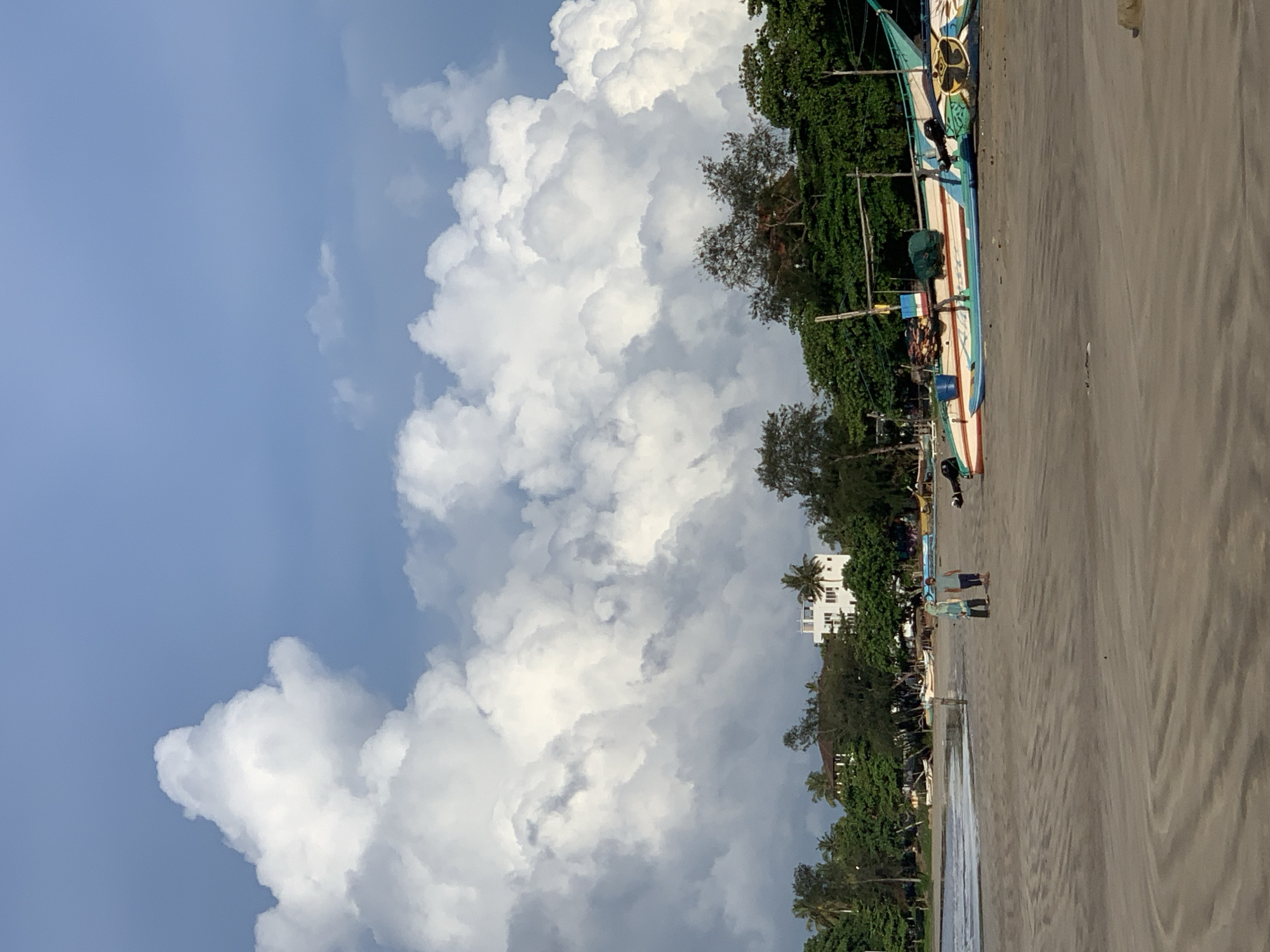 The image depicts a coastal scene in Weligama, Sri Lanka. The sky is predominantly blue, filled with massive cumulus clouds. The shoreline consists of a sandy beach. Several colorful outrigger fishing boats are pulled up onto the sand. Vegetation, including trees and shrubs, lines the land adjacent to the beach. People are walking along the shore. Buildings can be seen in the background.