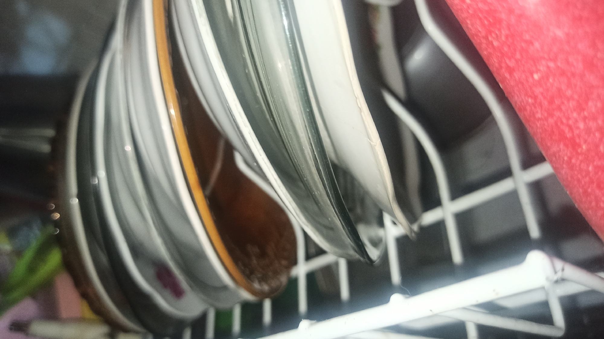 A stack of ceramic plates is visible in a dish rack. The plates are white with a thin brown or orange rim. The dish rack is made of white wire. In the foreground, a red object is partially visible. The scene appears to be indoors, possibly in a kitchen. The image does not provide any information about the city of Tuba, Indonesia, or any other location.