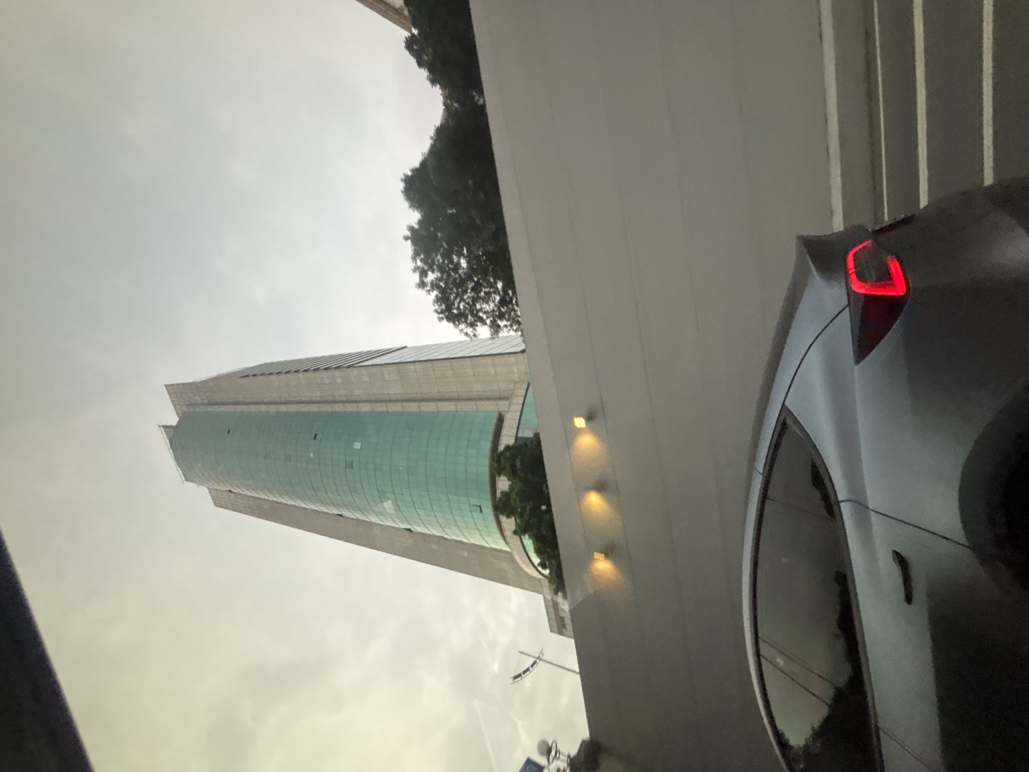 In Tianhe District, China, a silver Tesla Model 3 is captured from the rear passenger side, driving on a road. The car's brake lights are illuminated in red, indicating it may be braking or stopped. The car is positioned in the foreground, partially obscured by a gray concrete wall that extends horizontally across the middle of the frame. Behind the wall, a tall, modern skyscraper with a curved, teal-tinted glass facade dominates the background. The sky above is overcast and light gray, with some faint clouds visible. Lush green trees are visible at the base of the skyscraper and peeking over the wall. A street lamp or lighting fixture with a yellow glow is mounted on the wall. The overall scene suggests an urban environment, likely during daytime, with limited visibility due to cloudy weather.