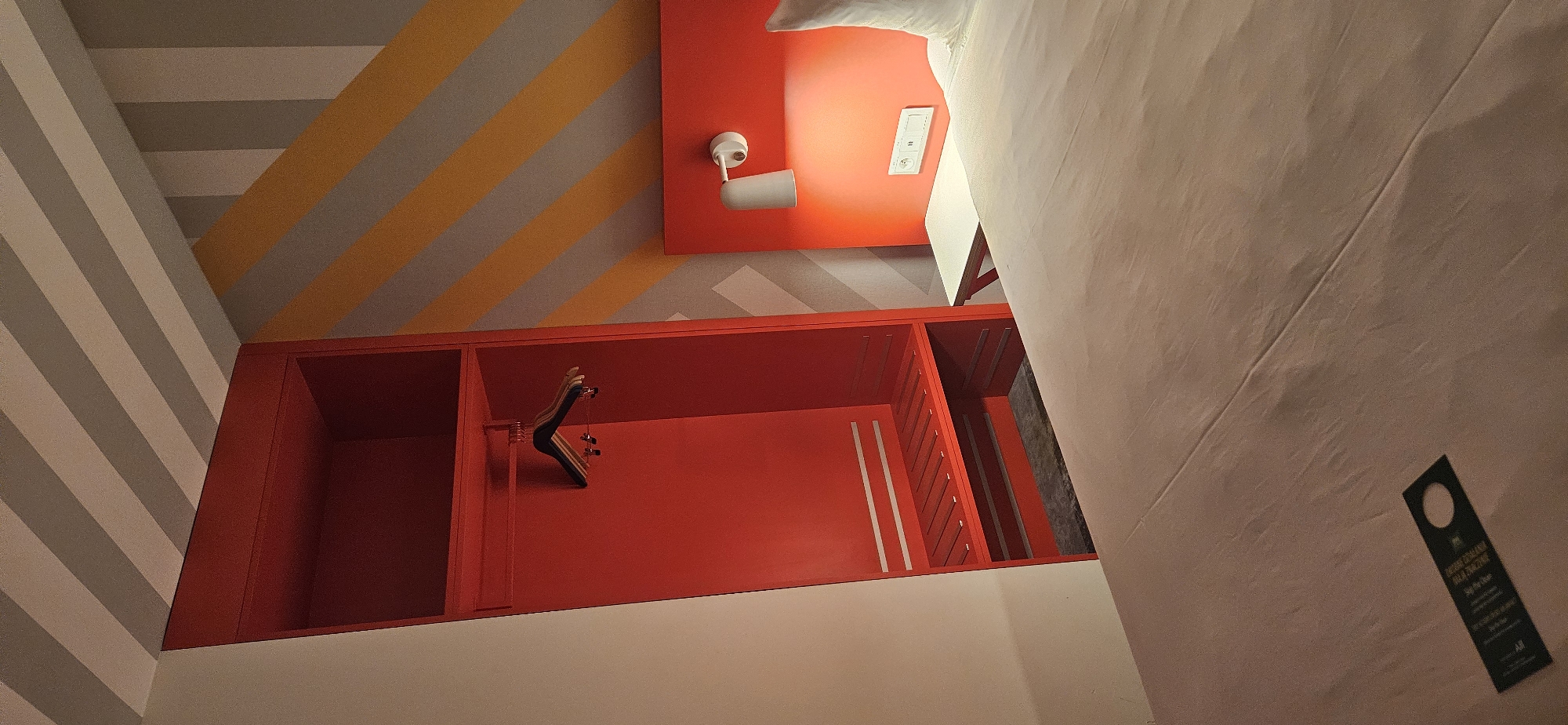 The image shows a modern hotel room in Warsaw, Poland. The room features a bright red wardrobe with empty hangers and a shelf. A bed with white linens is visible, with a red headboard and a bedside lamp. The walls are decorated with a geometric pattern of yellow and gray stripes. A do not disturb sign is placed on the bed. The overall impression is one of a clean and contemporary hotel room, likely designed for a business traveler or tourist.