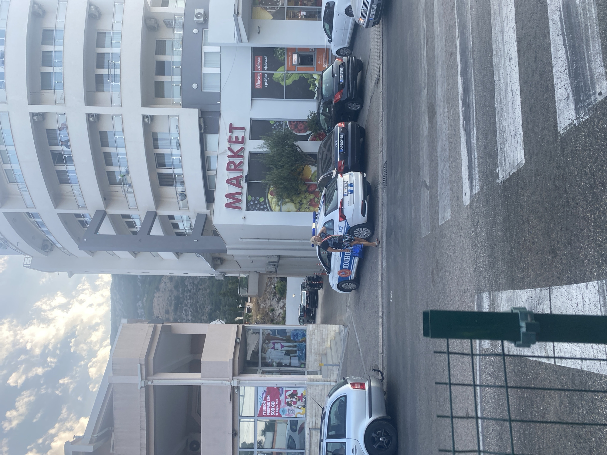 In Sutomore, Montenegro, a street scene unfolds with a modern, multi-story building dominating the right side of the frame. This building features a curved facade with numerous balconies. A sign above the ground floor reads "MARKET." To the left, a lighter-colored, less imposing building has a shopfront with displays of clothing, including swimwear. A promotional poster for "TELEKOM PREPAID SIM 500 GB" is visible in the shop window.
Several vehicles are present on the street. A white police car with blue flashing lights is parked, and a woman in a black top and patterned skirt stands near its rear door, holding a bag. A silver station wagon is parked in the foreground to the left, and other cars, including a black sedan and a dark hatchback, are parked behind the police car.
The foreground shows a crosswalk marked with white stripes on the asphalt. A dark green metal fence post is positioned in the lower left portion of the image, partially obscuring the crosswalk. The sky above is partly cloudy. The overall impression is an urban street scene on a bright day, with commercial activity and a visible police presence.