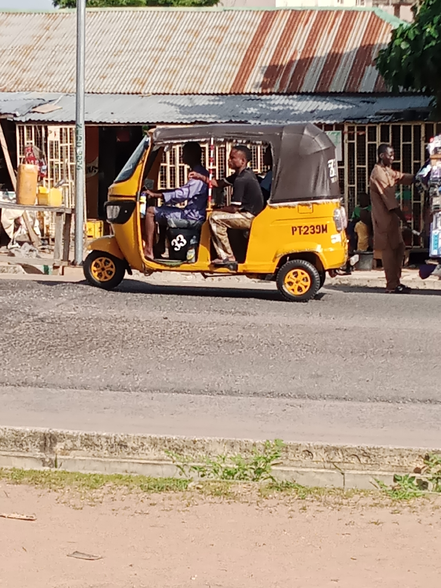 In Jalingo, Nigeria, a yellow auto-rickshaw, identified by the license plate "PT239M," is stopped on a paved road. The auto-rickshaw has a black canopy and carries two male passengers. The driver, wearing a blue shirt, is seated at the front. Behind him, a passenger in a black shirt is also seated. To the right of the auto-rickshaw, a man wearing a brown tunic stands near a storefront with barred windows, appearing to be interacting with a stall. The background features buildings with corrugated metal roofs and a visible light pole. A concrete curb and patchy vegetation line the side of the road in the foreground.