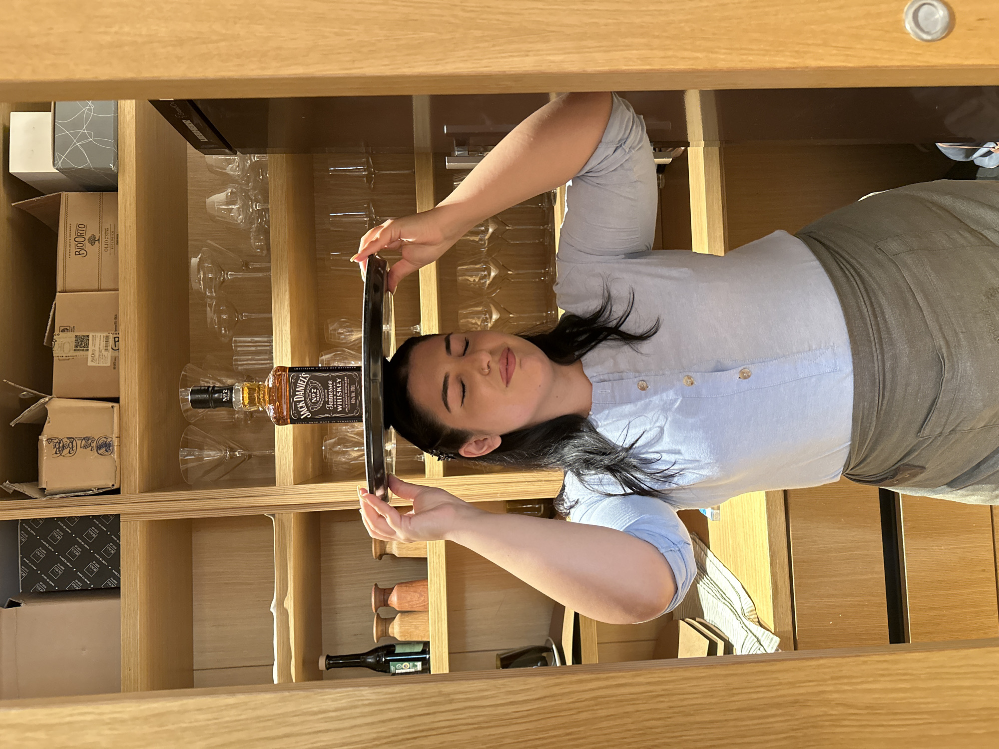 A young woman with dark hair, wearing a light blue button-down shirt and a grey apron, balances a tray with a bottle of Jack Daniel's Tennessee Whiskey on her head. She is standing in a wooden-paneled cabinet or pantry, likely in a restaurant or home setting. The shelves are stocked with various glassware, bottles, and boxes. The lighting is bright and appears to be artificial. The woman's eyes are closed, suggesting she is concentrating on maintaining her balance. The environment appears to be indoors, with a focus on organized storage. There is no indication of the city of Nicosia, Cyprus, in the image.