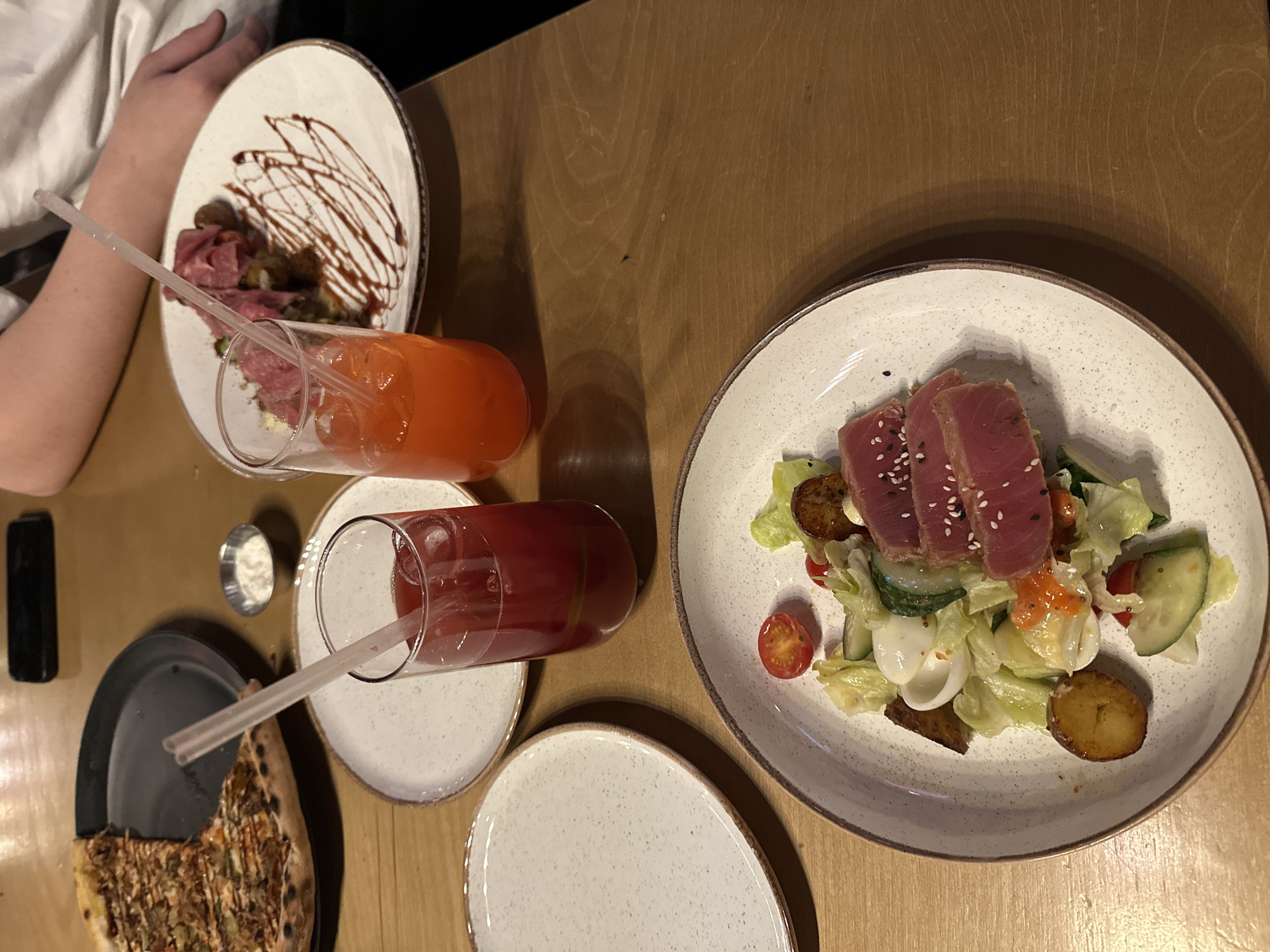 The image depicts a table at a restaurant in Izhevsk, Russia. The table is made of light brown wood. On the table, there are several dishes and drinks, including a plate with thinly sliced meat drizzled with dark sauce, a salad topped with seared tuna, a partially eaten pizza on a dark grey plate, and two tall glasses filled with red and orange drinks with straws. A small white container, potentially containing a sauce, is also present. A person's arm is visible in the upper left corner of the image.