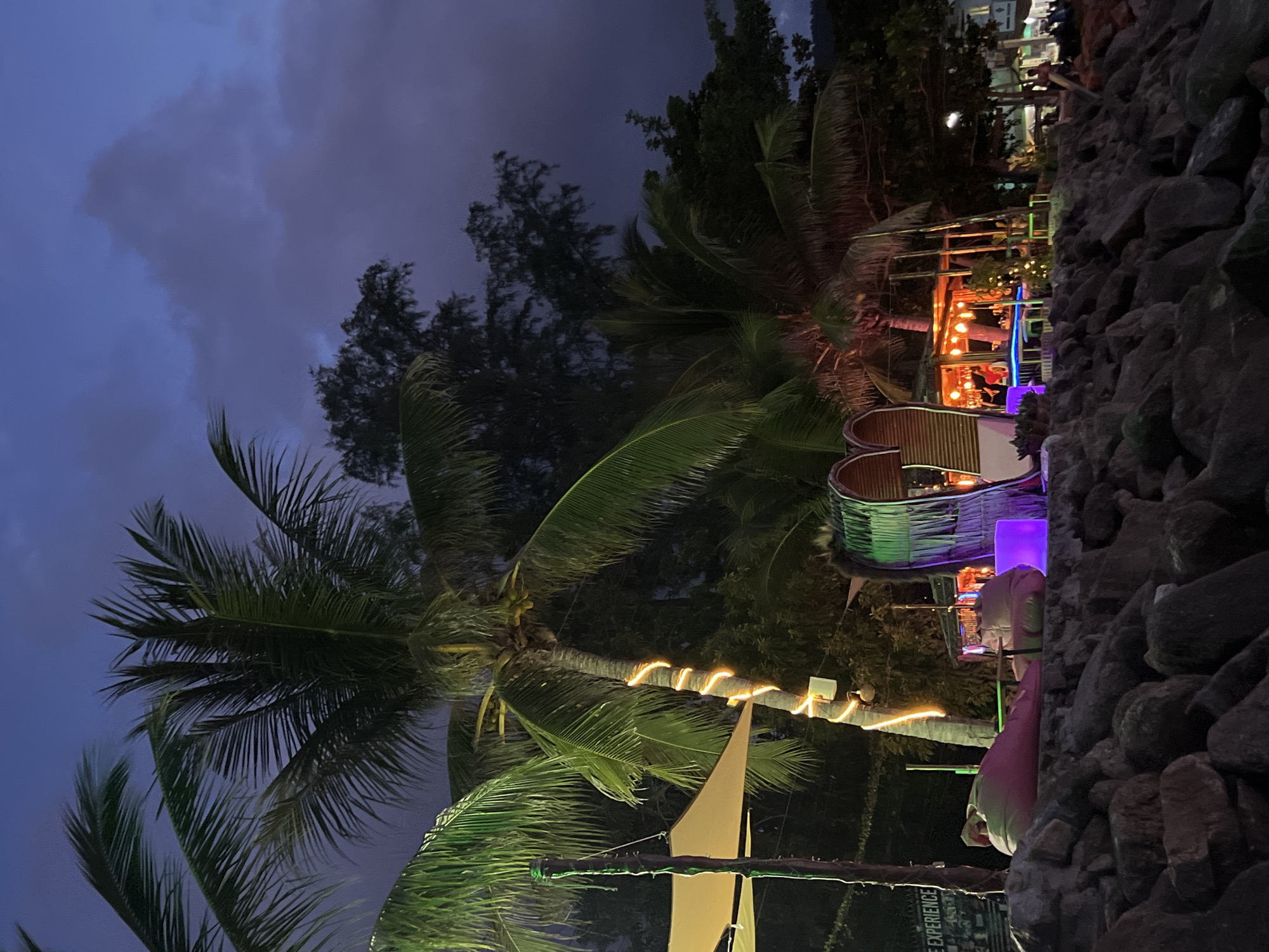 This image depicts a scene at dusk on the island of Ko Pha-ngan District, Thailand, likely a beachfront establishment. The foreground is dominated by a rocky embankment, with a pathway or seating area visible beyond it. Several beanbag chairs, one pink and one light green, are positioned on the seating area. To the left, a conical tent structure is partially visible, supported by poles. A prominent palm tree stands to the right of the tent, its trunk wrapped with warm-toned string lights. Behind the beanbag chairs and the palm tree, a structure with a heart-shaped entrance, adorned with a thatched roof and bamboo-like elements, is illuminated with purple and orange lights. Beyond this, a bar or restaurant area is visible, with tables and chairs set up and patrons present. More palm trees and dense foliage are interspersed throughout the scene. The sky above is a deep twilight blue, with visible clouds. In the distance, some buildings and lights suggest a populated area.