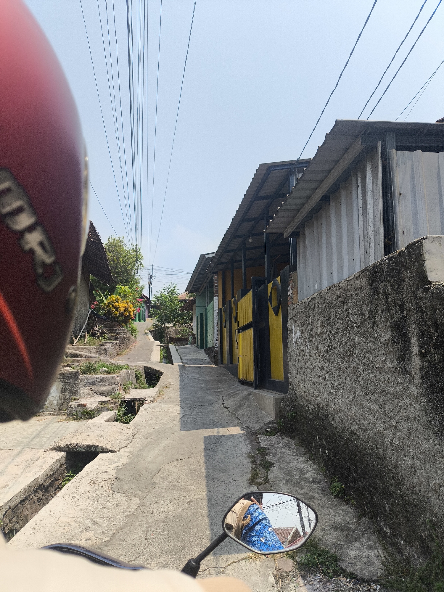 A point-of-view shot from a motorcycle helmet, looking forward down a narrow, paved street in Cilegon, Indonesia. The street is flanked by buildings and a concrete drainage system. On the left, a red helmet is partially visible in the foreground, with some of its branding showing. Beyond the helmet, a series of concrete steps leads up a small incline, overgrown with vegetation. In the distance, a path winds between buildings. On the right side of the street, a rough, textured concrete wall runs alongside a building with a corrugated metal exterior and a slanted, dark-colored roof. Yellow and black gates are visible behind the wall. In the motorcycle's rearview mirror, a reflection shows a person wearing a blue patterned garment and a backpack, with a rooftop visible in the background. Overhead, a dense network of electrical wires crisscrosses the sky. The day is bright and sunny, casting strong shadows.