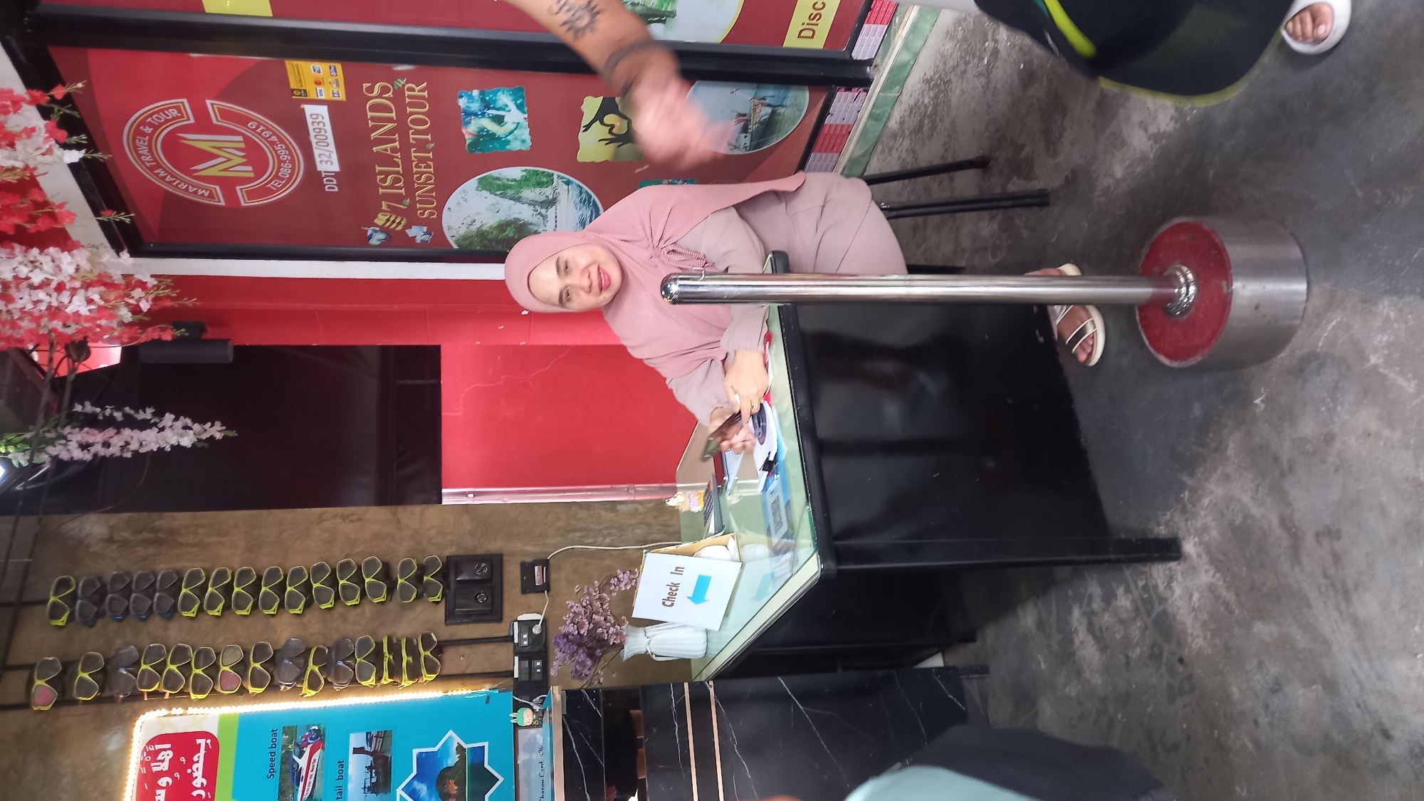 The image depicts the interior of a travel agency or tour operator, potentially in Ao Nam Mao, Thailand. A woman wearing a hijab is sitting behind a black counter with a glass top, working with papers or documents. On the counter is a "Check In" sign. Behind her is a sign advertising "7 Islands Sunset Tour" and another sign with the name "Mariam Travel & Tour". The wall displays several pairs of sunglasses and a promotional poster for boat tours. A decorative arrangement of white and red flowers is near the entrance of the tour operator. A large silver-colored metal barrier is to the right of the woman. The flooring is concrete. A person's arm with a tattoo is visible, and another person is partially seen wearing a black top.