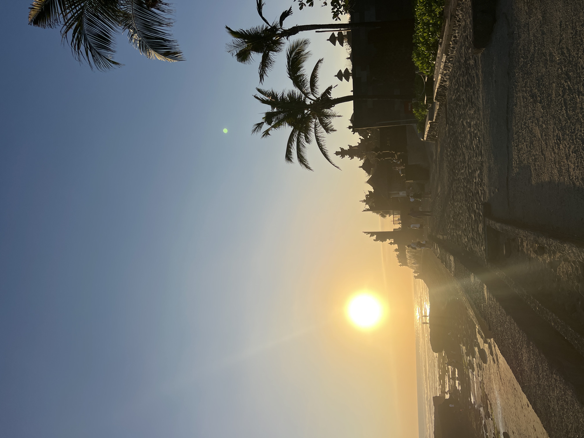 This is a photograph taken at sunset on a beach in North Kuta, Indonesia. The sun is low on the horizon, casting a warm golden light over the scene. The sky is a clear blue gradient, transitioning to a pale yellow near the sun.
In the foreground, there is a sandy path with textured footprints. To the left of the path, there is a stone wall and then a rocky coastline with the ocean. The ocean has gentle waves rolling in. In the mid-ground, there are several people walking along the path and on the beach. Some are silhouetted against the bright sun.
Further back, there are silhouetted structures that appear to be part of a temple complex, with tiered roofs. Two prominent palm trees with fronds are visible against the sky on the right side of the image. Lush green foliage is also visible along the side of the path near the temple structures. The overall atmosphere is serene and tranquil, capturing the beauty of a tropical sunset.