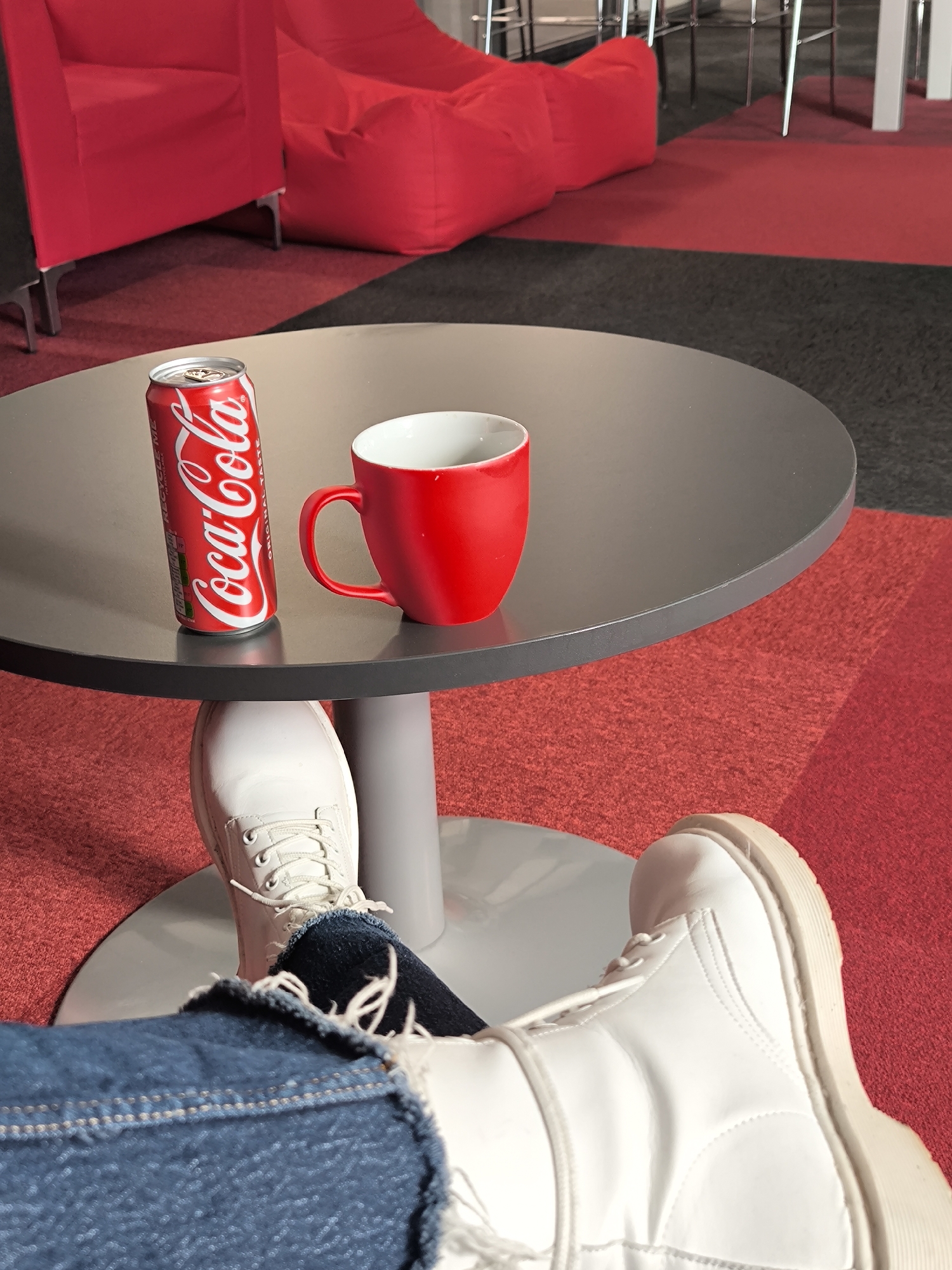 The image depicts a gray round table with a can of Coca-Cola and a red mug on it. Underneath the table, there are two feet wearing white boots and blue jeans. The floor is covered with a red carpet with black accents. In the background, there is a red armchair and a red beanbag chair. The location is unknown. The image focuses on the objects and attire, with no people visible other than the implied presence through the footwear.