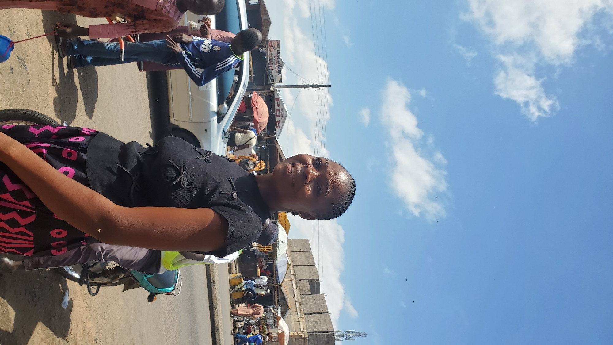 A medium shot shows a woman with dark skin and braided hair looking to her right, smiling. She is wearing a black short-sleeved shirt with decorative bows on the chest and skirt with a pink and black geometric pattern. She is standing on a street with other people and vehicles in the background. The environment appears to be a market or busy street in a city, with stalls, cars, and motorcycles visible. The sky is blue with some clouds.