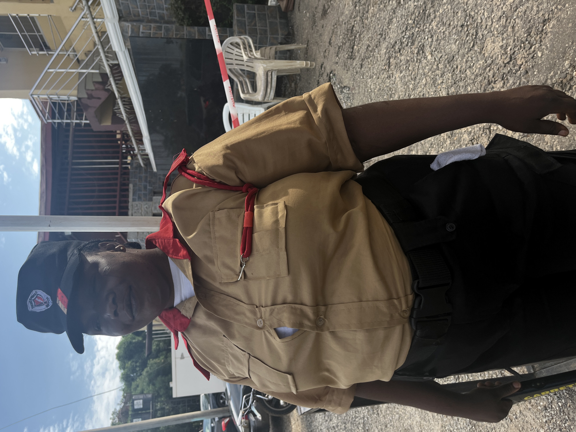The image displays an adult female security personnel standing outdoors in an urban setting in Abuja, Nigeria. She is positioned slightly angled to the viewer's left, looking forward. Her uniform includes a black cap featuring a red and white shield logo with the text "THUNDERBOLT SECURITY," a tan short-sleeved collared shirt with red trim on the collar and shoulder straps, and black trousers secured by a black tactical belt with a plastic buckle. The shirt has two buttoned breast pockets; one button is strained, revealing a white undershirt. A red lanyard is attached to her right breast pocket, and a small white cloth is tucked into her right hip pocket. She is holding a yellow and black Garrett metal detector in her left hand.
The background consists of a light-colored building with an external staircase featuring metal railings on the right. Below the building is a dark, tiled wall and a reflective surface, possibly a gate. A red and white striped barrier tape is strung horizontally across the mid-ground, with partially legible text. Several plastic chairs, including one white and others off-white, are stacked and placed to the far right. The ground is an unpaved, gravelly surface. In the far left background, partial views of a motorcycle, a car, and green foliage are discernible, along with a structure resembling a water tank on a stand. The sky above is blue with scattered white clouds, indicating bright daylight conditions.