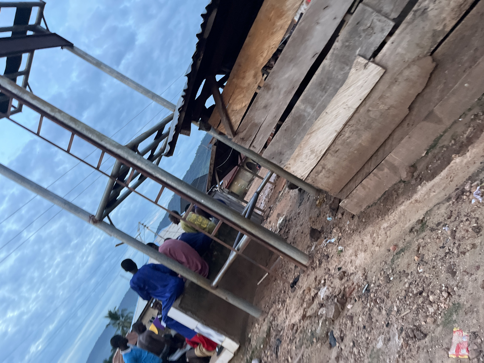 An outdoor scene shows a group of people, likely male, sitting on a raised concrete structure. They are dressed in traditional attire, including a blue and pink robe. The environment appears to be a developing or rural area, with construction materials, including wooden planks and metal scaffolding, visible. Some of the people are facing away from the camera, while others are partially obscured. In the background, there are some buildings, vegetation, and hills. The sky is overcast with a cloudy, blue hue. The ground is rough and unpaved, with scattered debris.