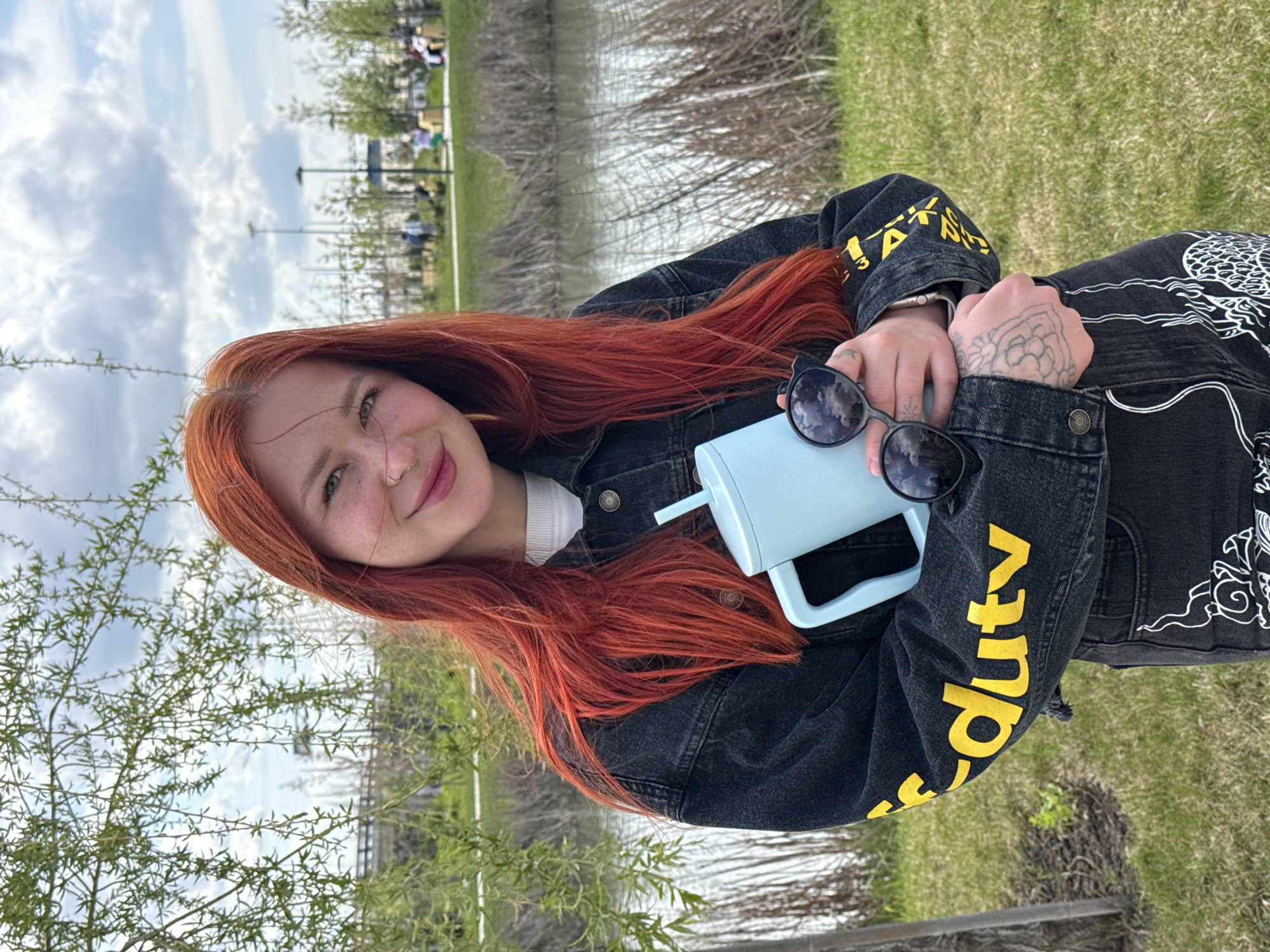 A young woman with red hair stands outdoors next to a body of water, likely a lake or river, possibly in a park. She is wearing a black denim jacket with yellow lettering and a white dragon design. She holds a light blue water bottle and a pair of sunglasses in her hands. Her left hand has a tattoo on it. The background shows green grass and trees. The location is Rostov-on-Don, Russia.