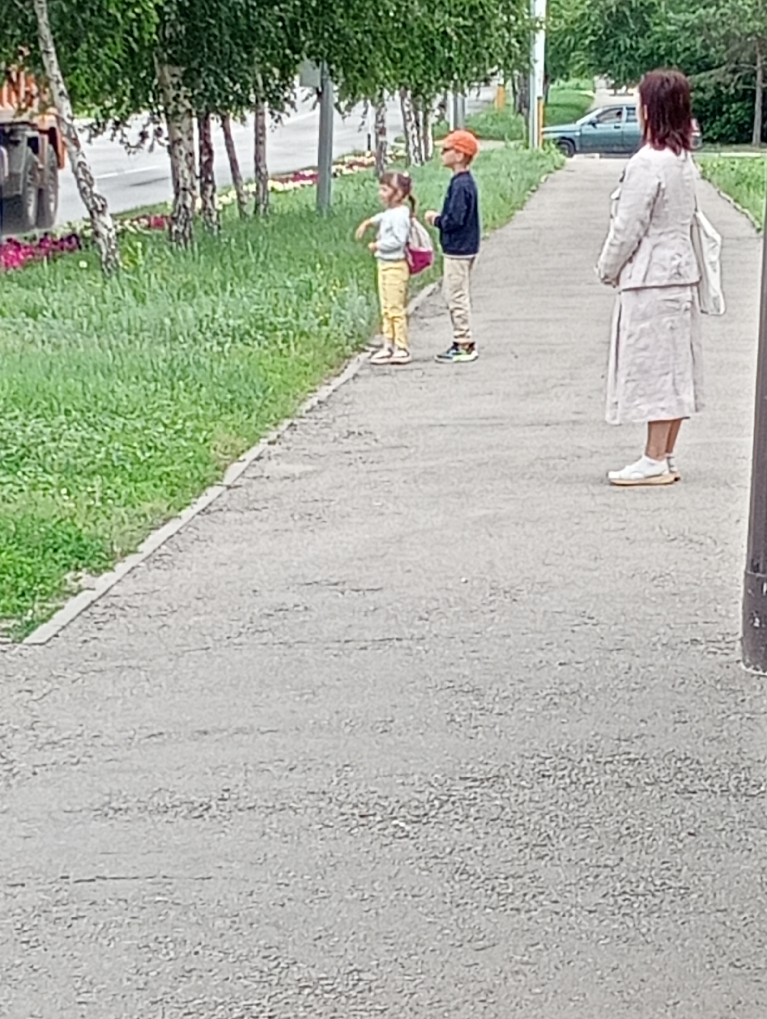 A medium-shot, eye-level, full-color, outdoor photograph shows a paved walkway next to a grassy area with trees. In the foreground, a woman stands facing away from the camera, looking to the left. She is wearing a light-colored trench coat and a long skirt, with white sneakers on her feet. Behind her, two children stand facing the same direction. A girl, wearing a light-colored shirt and yellow pants, has a pink backpack on her back. A boy, wearing an orange baseball cap and a dark jacket, stands next to her. In the background, a dark-colored vehicle is visible on a road. The scene appears to be in a park or residential area with trees and grass lining the walkway.