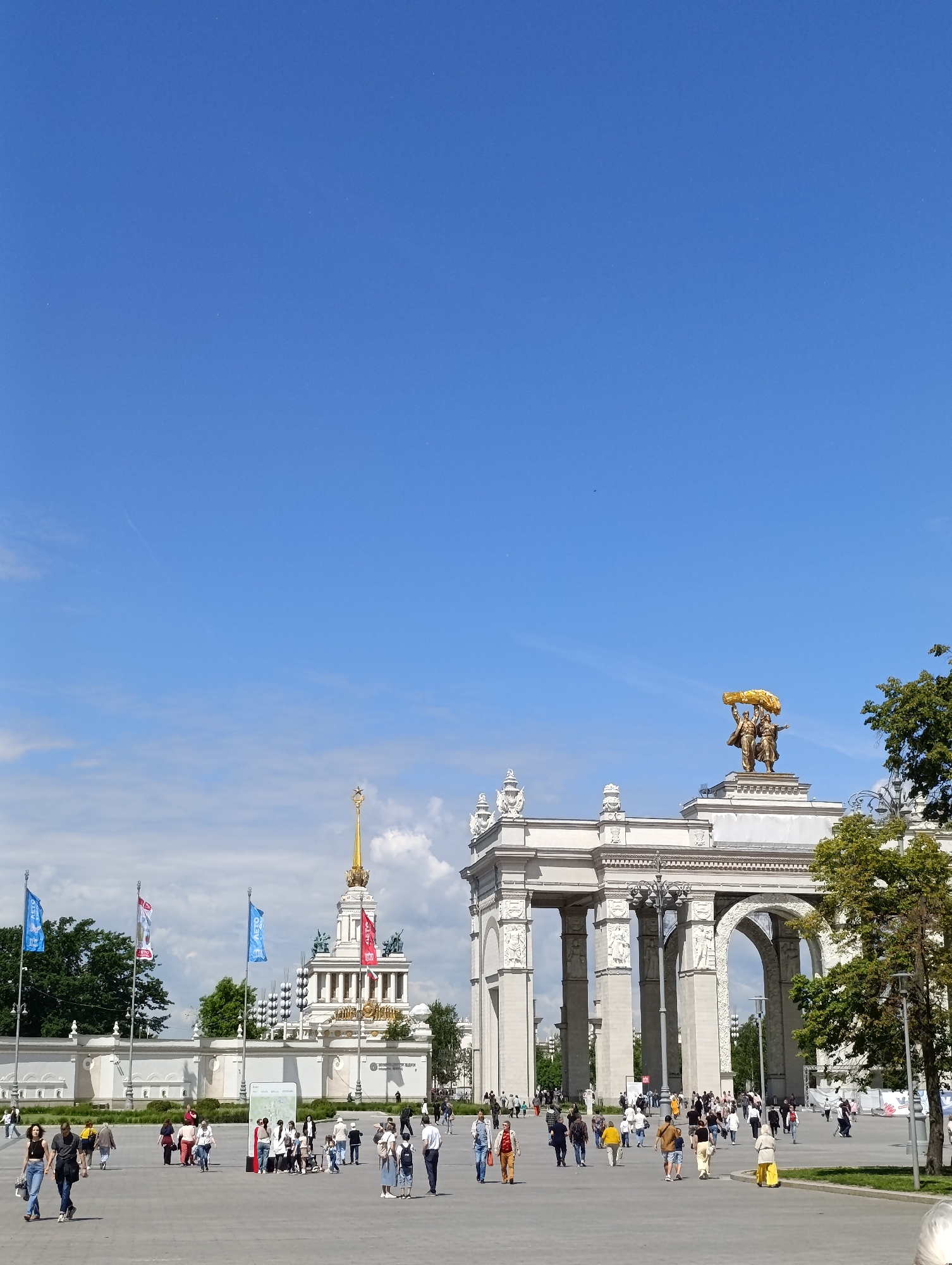 This brightly lit photograph captures a bustling scene at a grand architectural complex, identified as the Exhibition of Achievements of National Economy (VDNKh) in Moscow, Russia. The image is taken on a sunny day under a clear blue sky with a few wispy clouds.
In the foreground and mid-ground, numerous people are seen walking across a vast, paved plaza. They are casually dressed, suggesting visitors or tourists enjoying the outdoor space.
The right side of the frame is dominated by a majestic, light-colored triumphal arch, adorned with ornate classical carvings. Crowning this arch is a prominent golden sculpture depicting two figures, a man and a woman, holding a large golden sheaf. This is the Main Entrance Arch of VDNKh, featuring the "Tractor Driver and Kolkhoz Woman" sculpture.
Further back on the left, a tall, multi-tiered pavilion stands out with its white facade and a distinctive golden spire topped with a red star, characteristic of Soviet-era architecture. This is likely the Central Pavilion of VDNKh. Several blue flags with "АВТО" (AUTO) visible in white text are displayed on poles in front of this pavilion. Green trees are visible on the far right and in the background, adding foliage to the monumental setting. The overall scene conveys a lively atmosphere of public leisure and visitation at an iconic Moscow landmark.