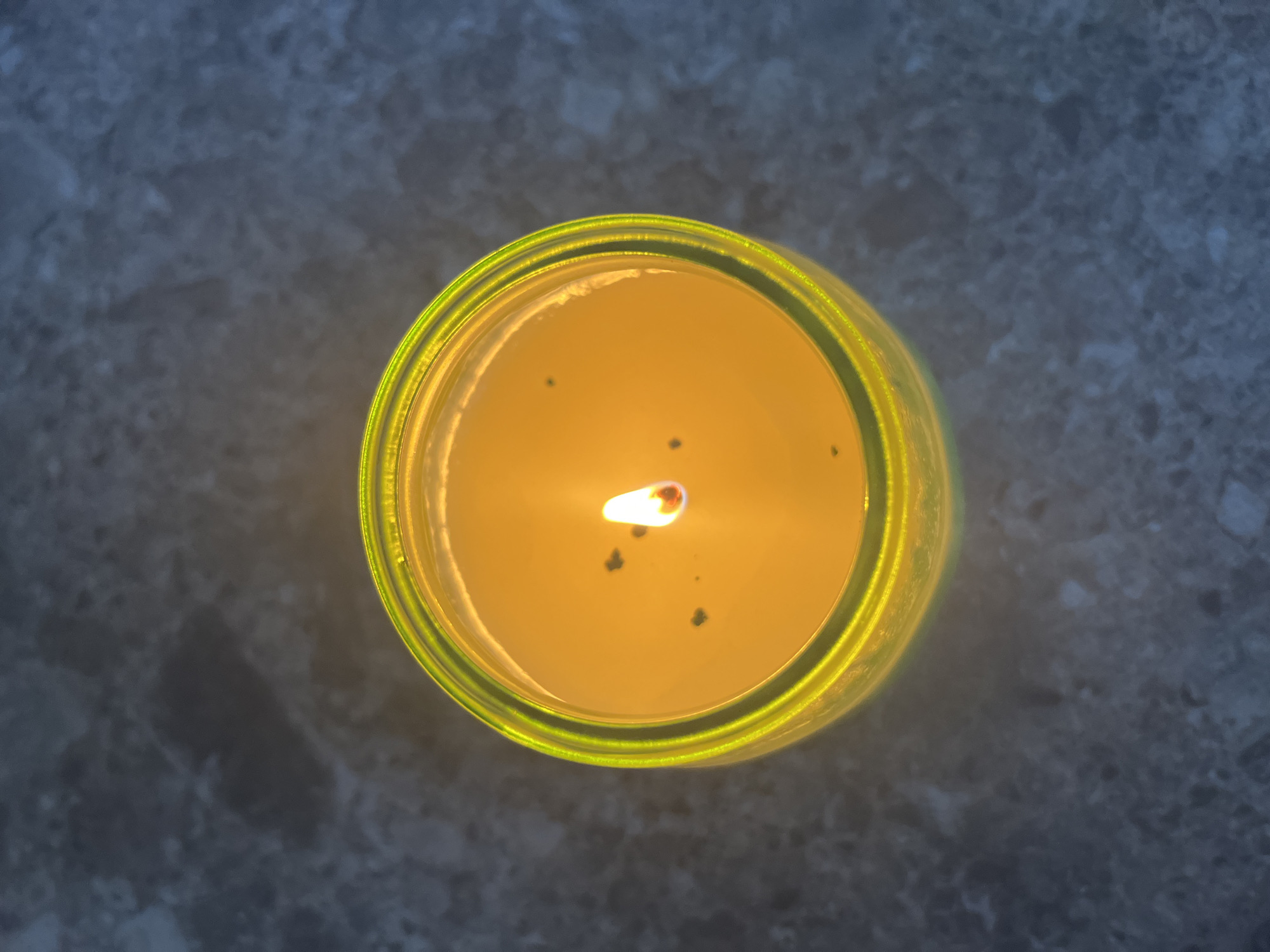 A lit candle is positioned on a gray, stone-like surface. The candle is inside a green glass container and the wick is burning. Several black specks are present on the candle's surface. The location is unknown.