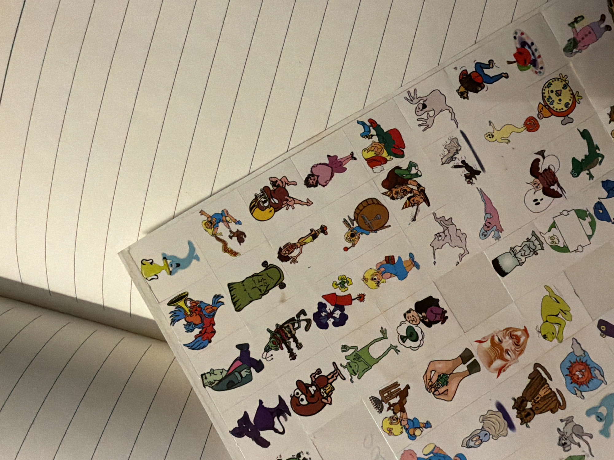 The image depicts a page of stickers, likely from a sticker book, placed on a notebook. The stickers are cartoonish and feature a variety of characters and objects. The notebook is open and shows lined pages. The location and city are unknown.