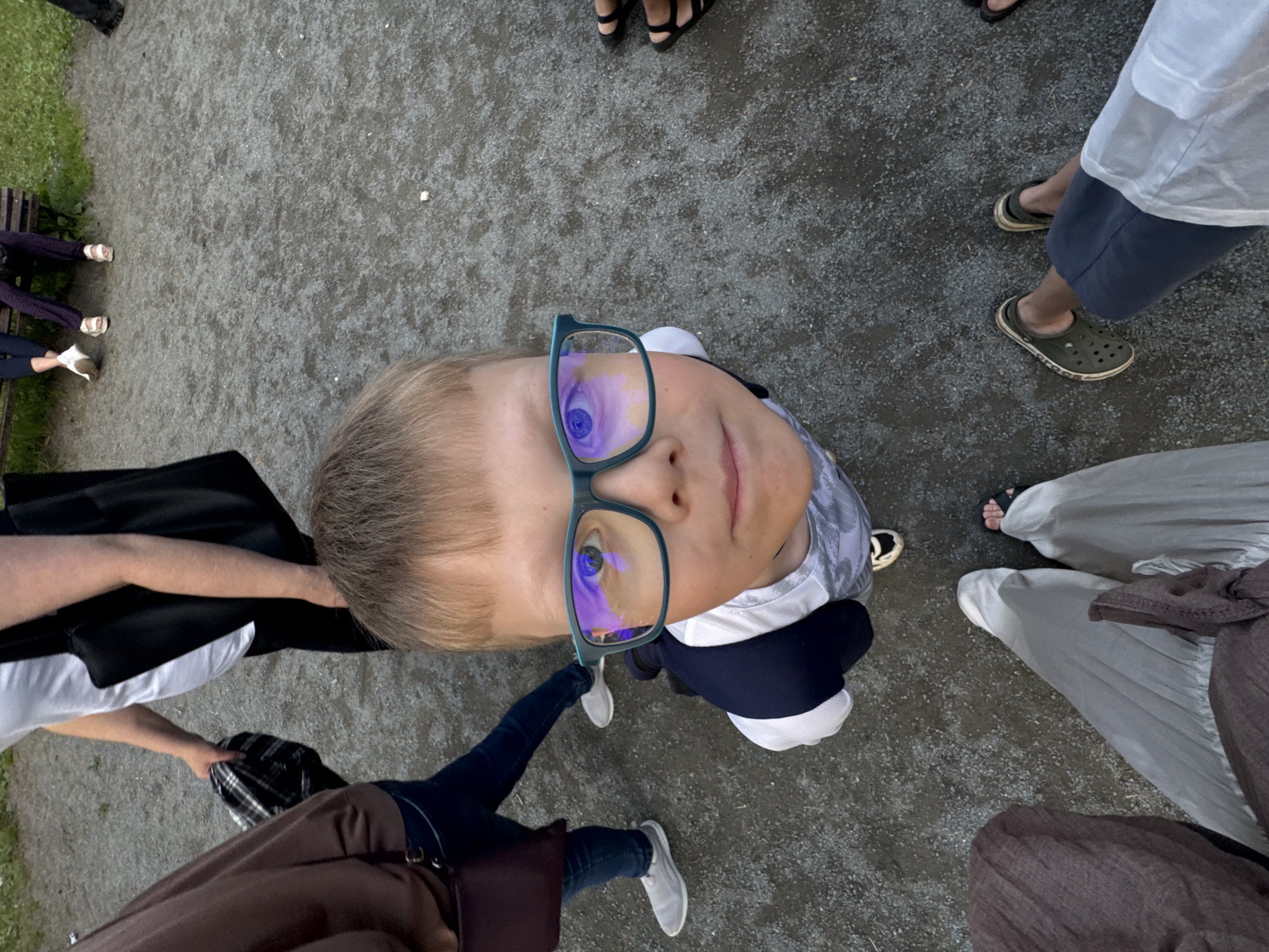 A group of people are standing and sitting in a park-like area in Горный, Russia. In the foreground, a young boy wearing glasses looks up at the camera. He is wearing a dark blue jacket over a white t-shirt. Behind him, other people are visible, some standing and some sitting on a bench. The ground is covered with gravel. The overall scene suggests a casual outdoor gathering.