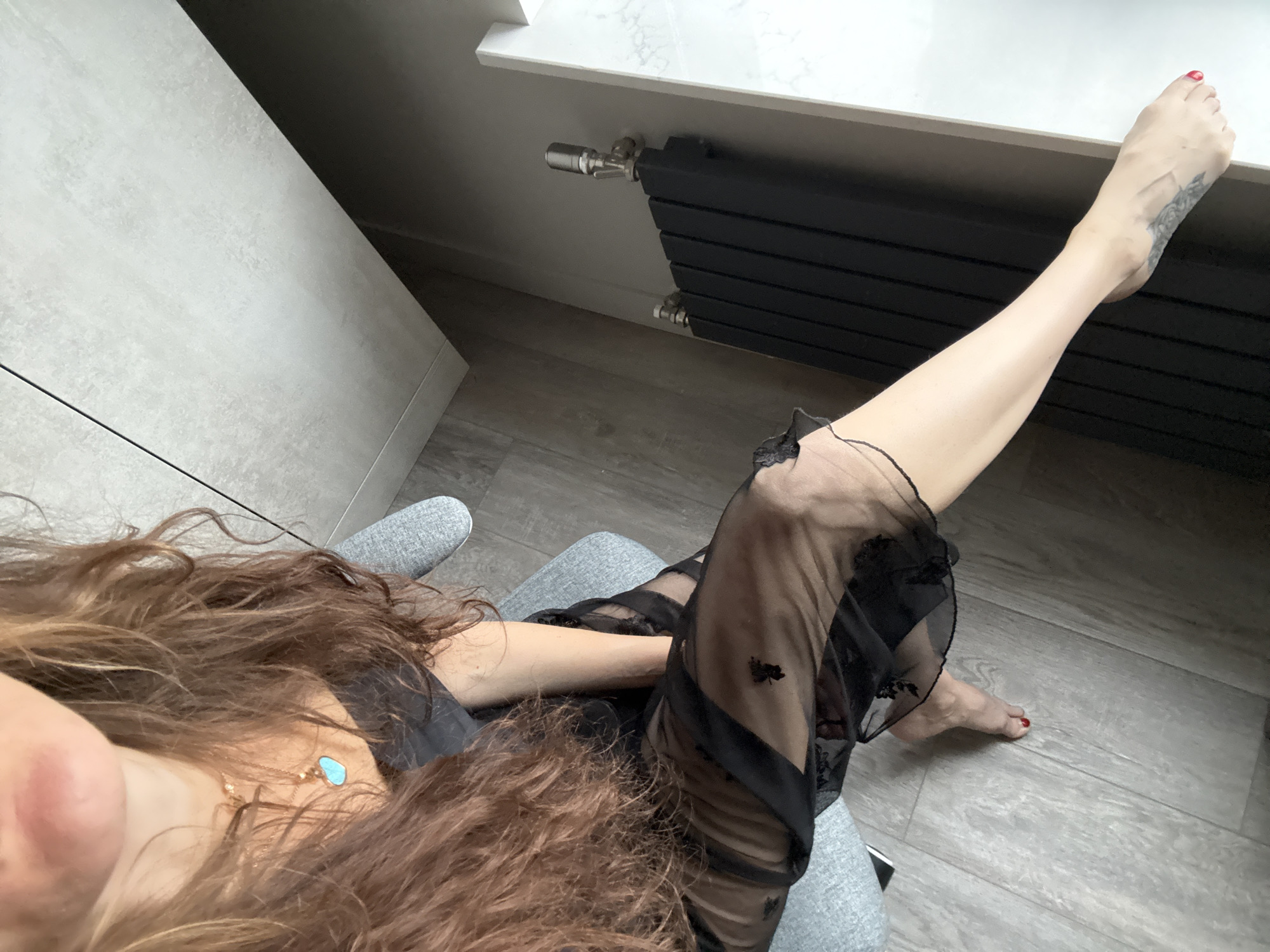The image shows a person sitting in a chair, presumably indoors. The person's left leg is extended upwards, with the foot near a black radiator. The person is wearing a black, semi-sheer garment. The person has long, brown, wavy hair. The person has a necklace with a turquoise pendant. The background includes a grey wall and a grey floor. The location cannot be determined to be Khimki, Russia based on the visual information provided.