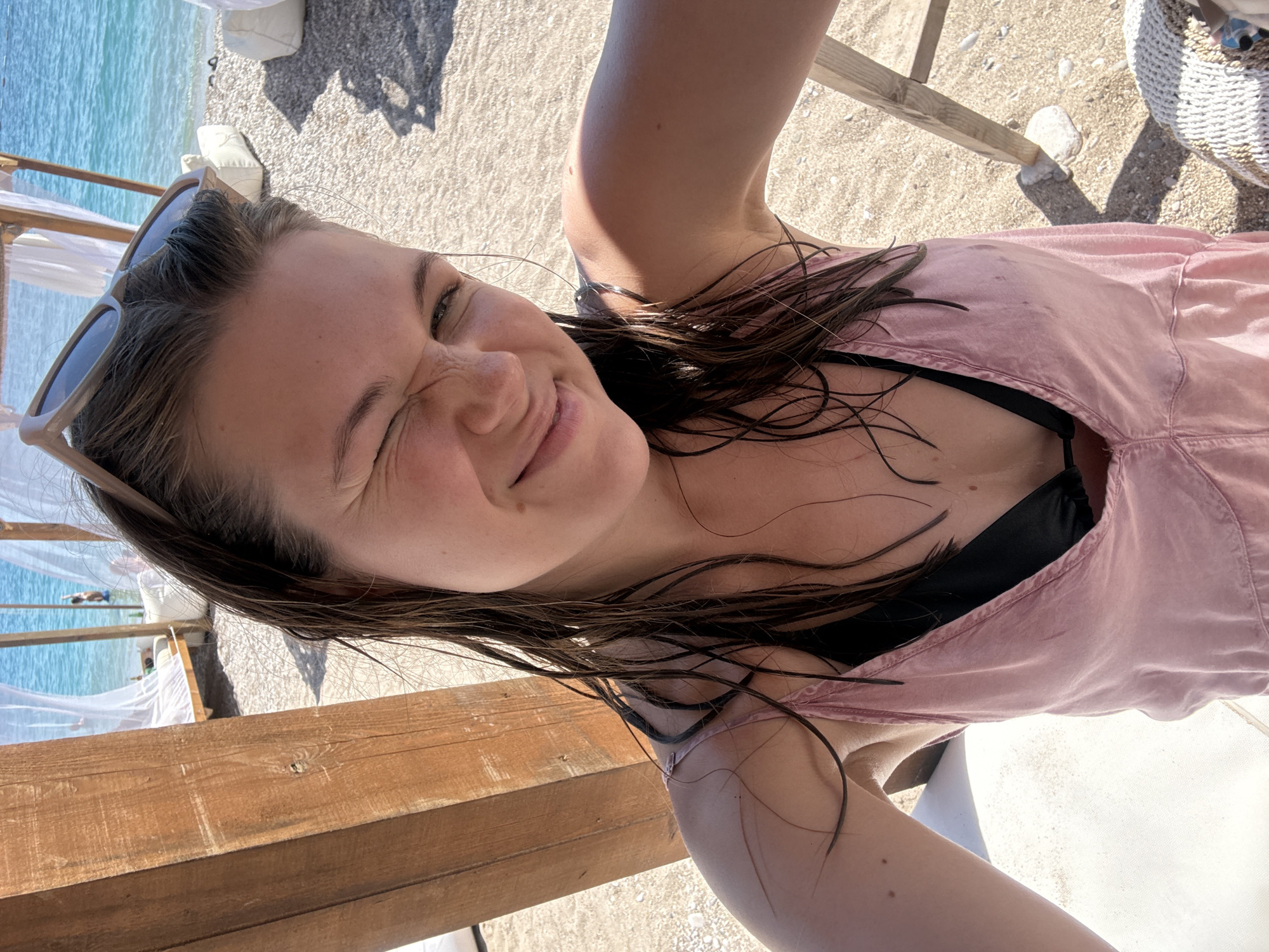 A young adult female is captured in a close-up selfie on a sunny beach in Drobnići, Montenegro. She is winking with her right eye and smiling slightly, with wet, dark brown hair pushed back, adorned with light-colored sunglasses on her head. She wears a pink, sleeveless, loose-fitting top or dress over a black bikini top. Her left arm, tanned with a few small moles visible, extends towards the camera. To her left, a sturdy wooden post of a beach structure is prominent. The background reveals a pebbly beach leading to clear, turquoise blue water, where several white, draped beach cabanas are set up. One person is visible standing in the shallow water. White, beanbag-style seating is scattered on the sand, and the bottom right corner shows a segment of a light-colored woven bag or basket. The scene is brightly lit by direct sunlight, casting distinct shadows on the sand and wooden elements.