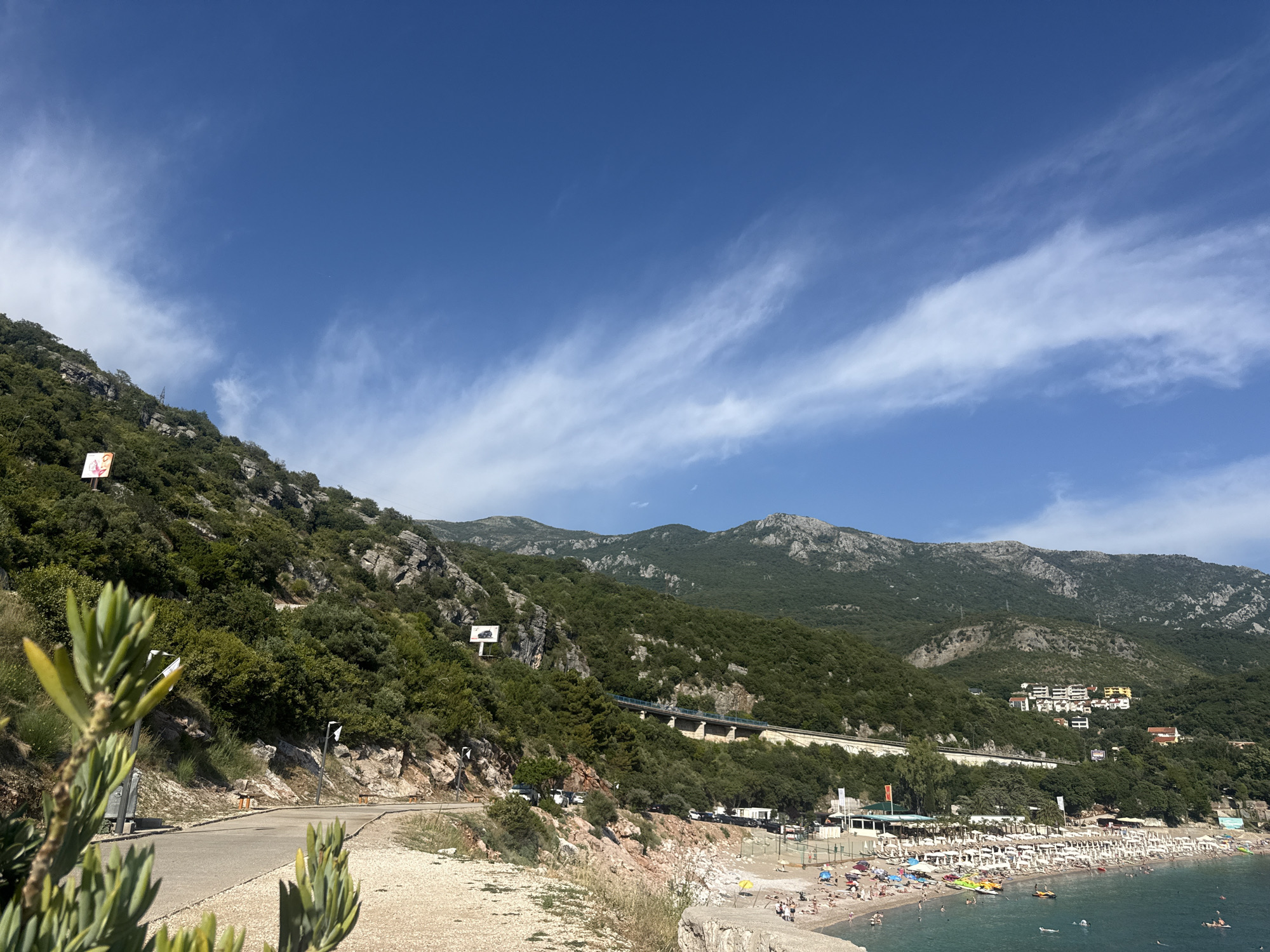 The image captures a scenic view of the Kamenovo, Montenegro coastline on a clear day. In the foreground, a paved road winds along the side of a lush, green, rocky hillside, with a few sparse plants in the immediate foreground. The hillside is covered in dense vegetation and dotted with some exposed rock formations. Several signs are visible on the hillside, including a billboard with a car advertisement.
Further down, the road appears to lead to a bustling beach area. The beach is lined with numerous white umbrellas and lounge chairs, indicating a popular tourist destination. People are scattered across the beach, some sunbathing, others walking, and some are in the clear turquoise water, swimming or enjoying water activities. A few boats and watercraft are also visible in the sea.
In the background, the imposing silhouette of mountains rises, covered in greenery. A section of elevated highway or infrastructure can be seen traversing the mountainside, partially obscured by trees. Several buildings, including apartment complexes and smaller structures, are nestled into the slopes of the mountains overlooking the bay. The sky above is a vibrant blue, with wisps of white clouds streaking across it, suggesting pleasant weather. The overall scene depicts a lively coastal resort town integrated into a dramatic natural landscape.