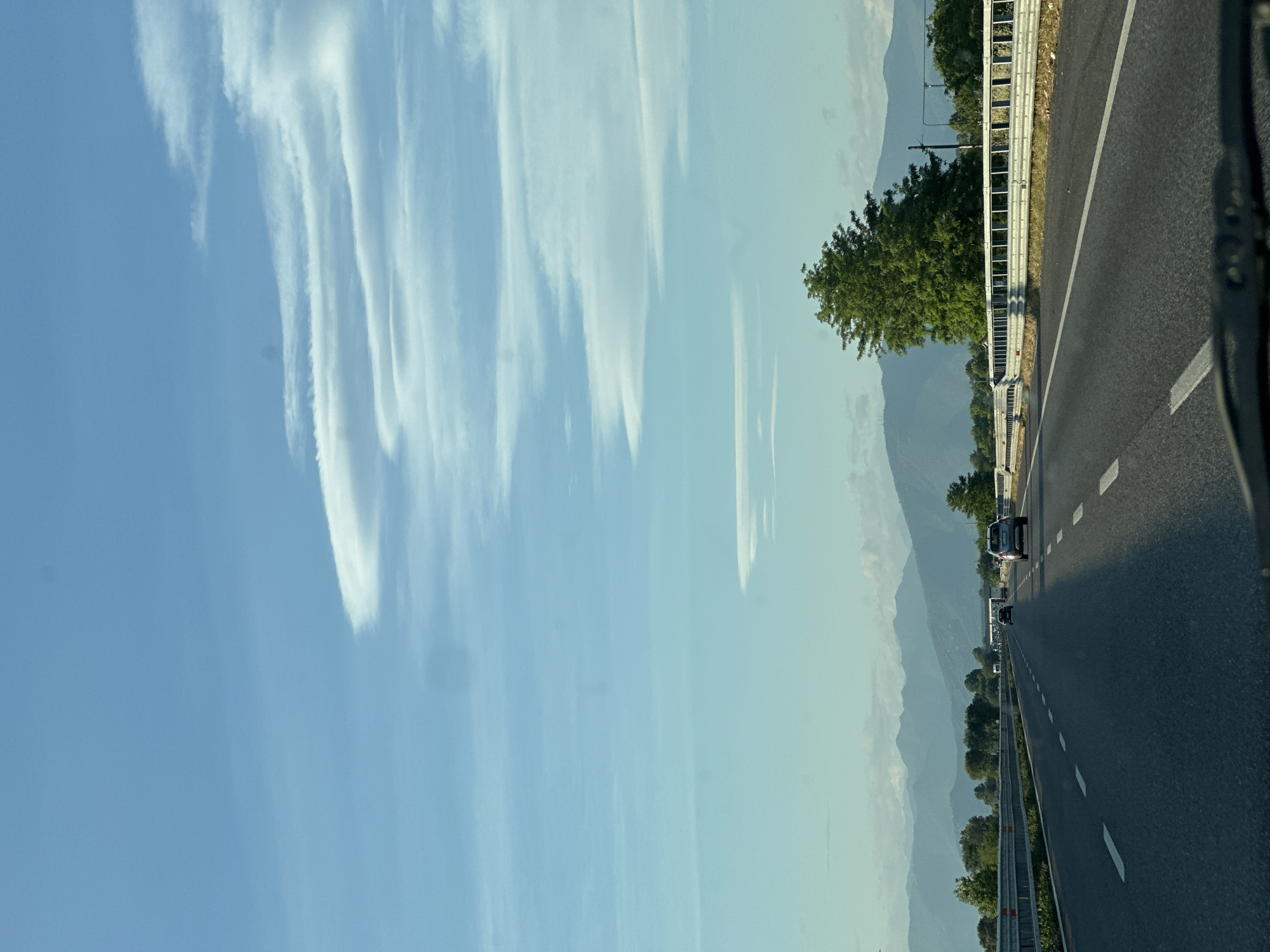 An asphalt highway stretches towards distant mountains under a bright blue sky dotted with wispy white clouds. The highway has two lanes in each direction, separated by a solid white line, and a dashed white line dividing lanes within each direction. Several vehicles are visible on the road, including a silver car in the right lane and a dark-colored car in the left lane. On the right side of the highway, a metallic guardrail runs parallel to the road, with a lush green tree standing tall behind it. The background features a range of hazy, blue-toned mountains. The image is captured from a vehicle traveling on the highway, with the dashboard visible at the bottom. The location is identified as the city of Leno, Italy.