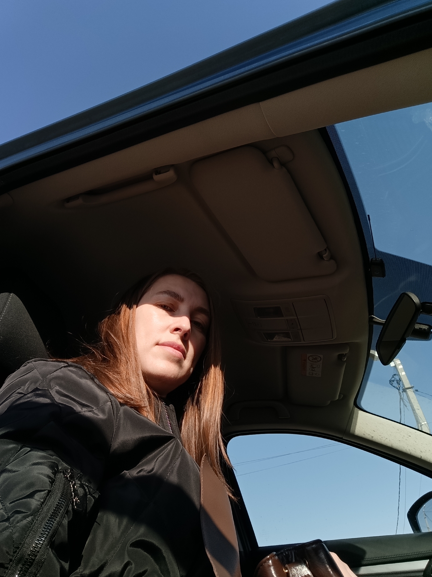 A medium shot depicts a woman inside a car on a sunny day. The angle suggests she may be in the driver's seat, but her hands are not visible on the steering wheel. She is wearing a black quilted jacket and a seatbelt across her chest. Light falls on her face and hair, which is brown and shoulder-length. The interior of the car, including the rearview mirror, sun visor, and roof lining, are visible. The car window reveals a clear blue sky and power lines in the background. The photograph might have been taken in the city of Chaikovsky, Russia, but there's no clear visual indication within the image itself to confirm this location.