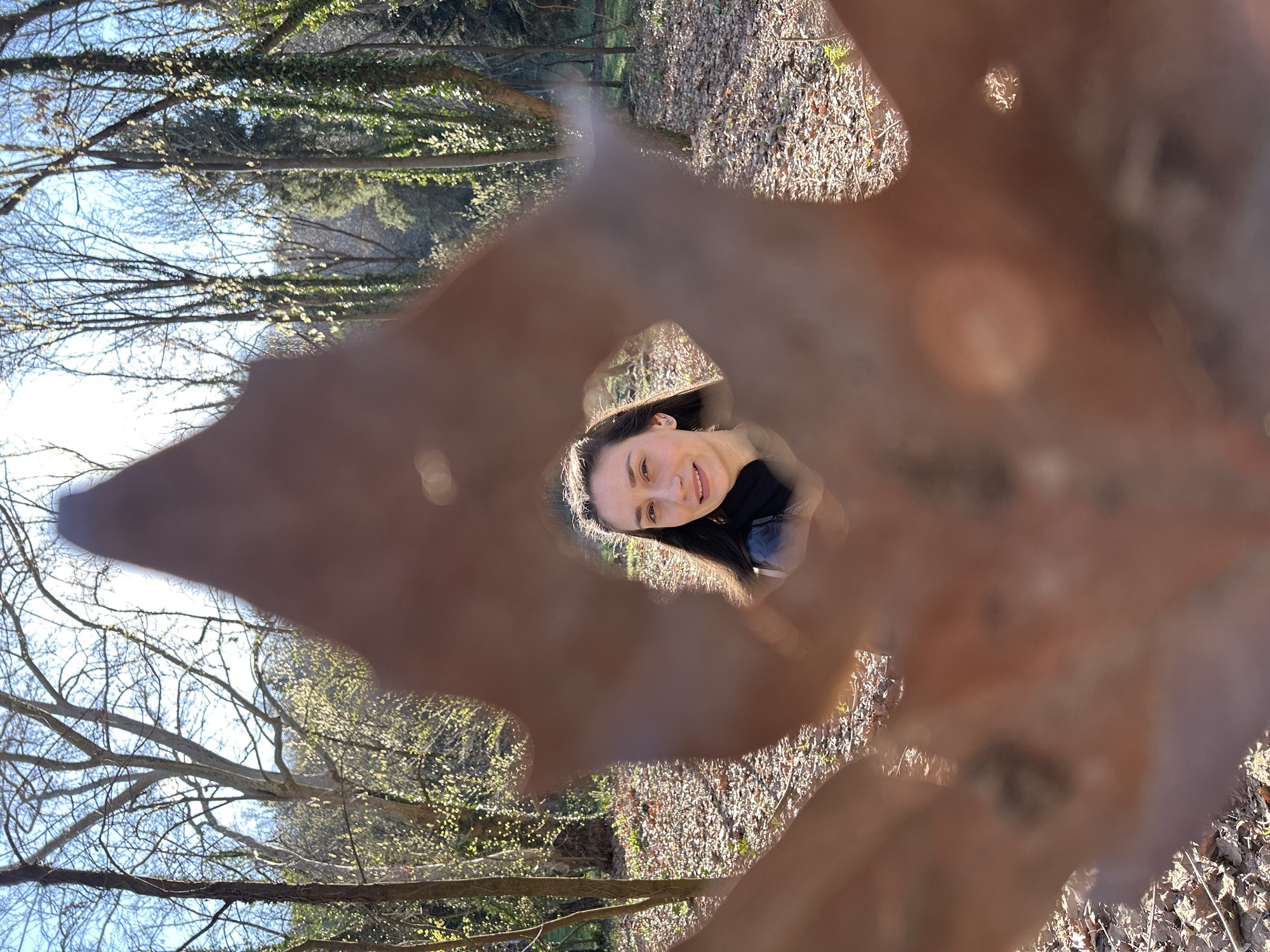 The image shows a woman's face framed by a large, brown leaf with a hole in it. The background appears to be a wooded area with bare trees and leaf-covered ground. Based on the location provided, the scene is possibly in a park or forest in Pleven, Bulgaria.
