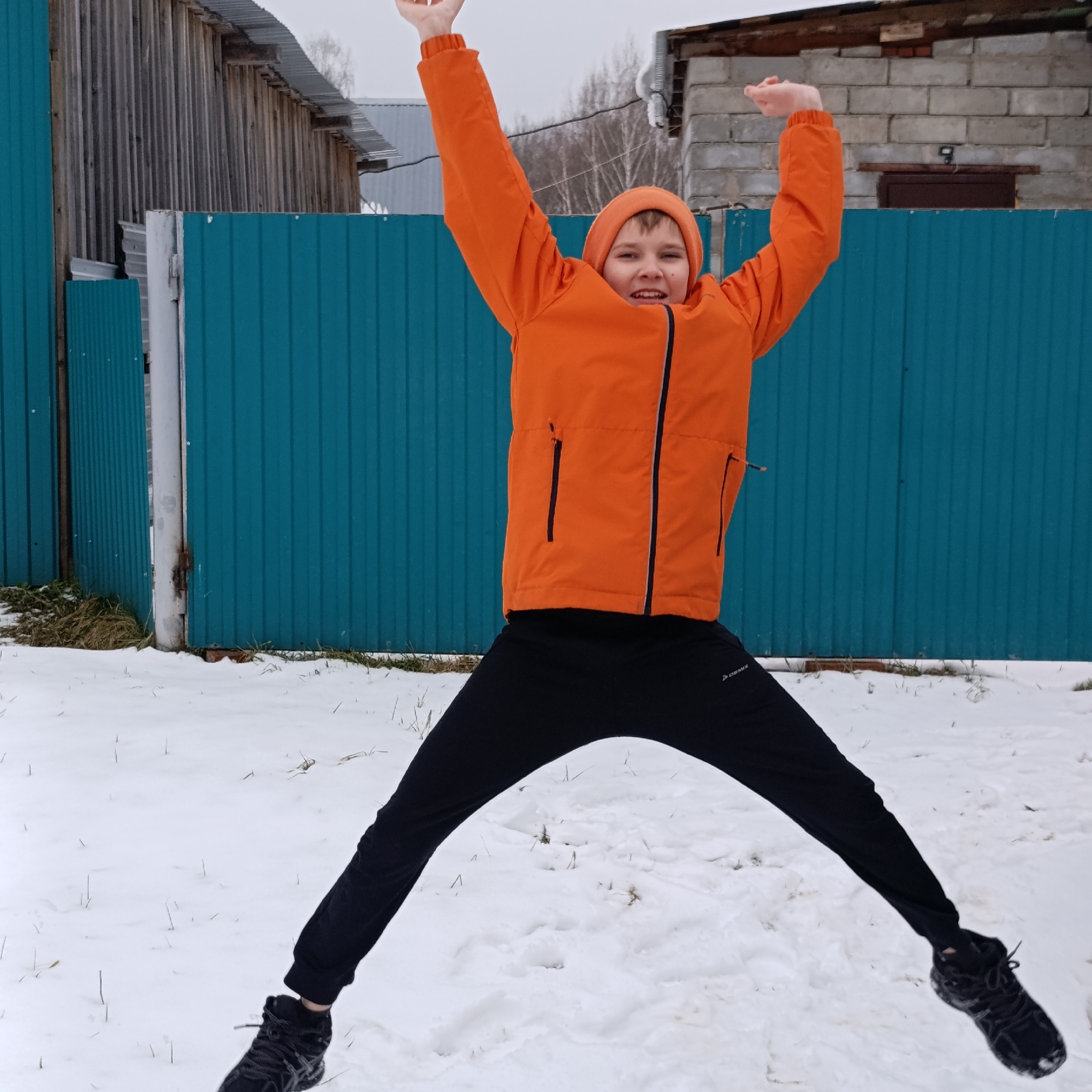 A young person jumps in the air in a snowy, outdoor environment. The individual is wearing an orange hooded jacket and black athletic pants. They have both arms raised above their head and their legs extended outward. Behind them is a bright blue corrugated metal fence. A wooden building and a brick structure are visible in the background. The setting is a snowy area with sparse vegetation visible through the snow. The location is Dubovaya, Russia.