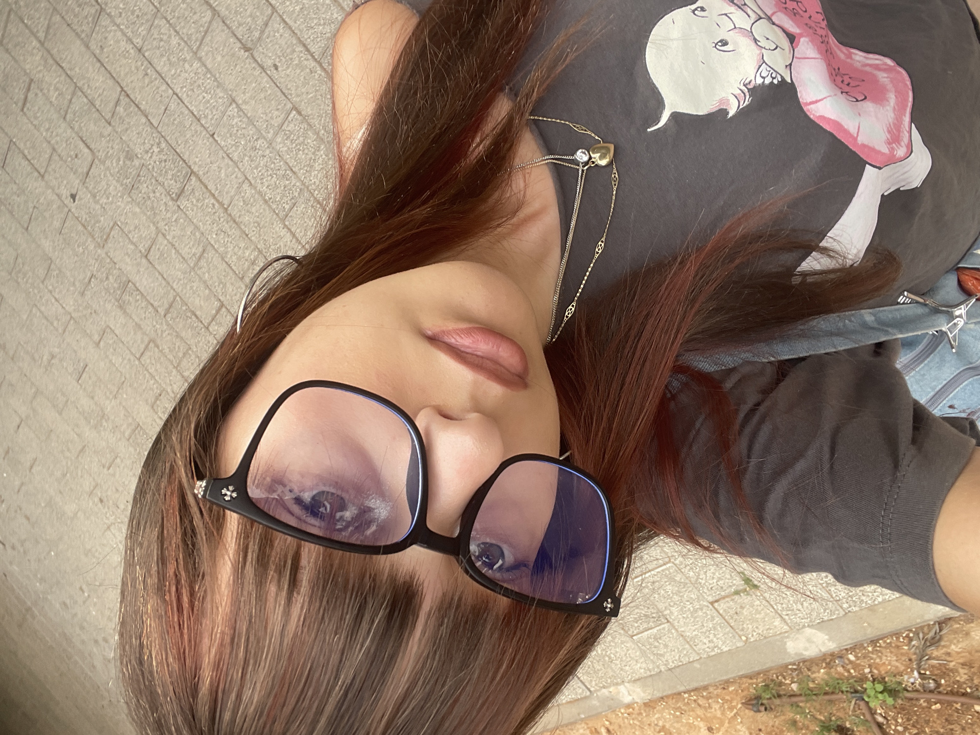 The image is a selfie taken by a person with long, reddish-brown hair, wearing glasses and a dark grey t-shirt with a graphic of a cartoon baby on it. The person is looking towards the camera, with their face angled to the left. The background appears to be a paved walkway, possibly a sidewalk or pedestrian path, with visible cracks and a repeating brick pattern. To the left of the walkway, there is an area with dry soil and some sparse green vegetation, suggesting an outdoor environment. The lighting indicates it is daytime, likely outdoors. While the specific location is not visible, the question asks to include "the location in the city of Raanana, Israel," which is information external to the image content itself. Therefore, assuming this context is provided, the scene is of a person taking a selfie outdoors in Raanana, Israel, on a paved walkway.