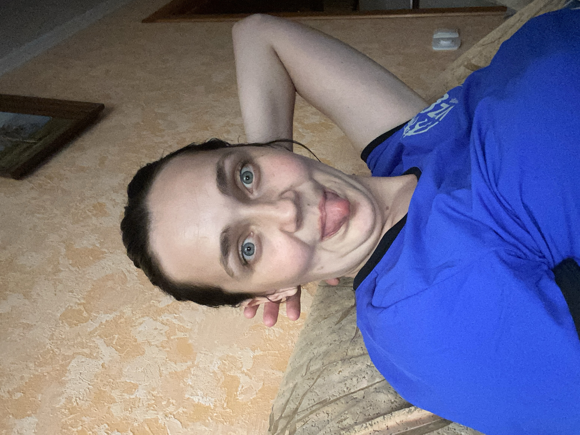 In an indoor setting, a Caucasian woman with dark hair pulled back in a bun and blue eyes makes a humorous facial expression with her tongue sticking out. She is wearing a bright blue t-shirt with a black trim around the neckline and a logo on the left sleeve. Her right arm is bent behind her head. The background features a textured, light orange wall and a portion of a couch with a patterned fabric. A framed picture hangs on the wall to the upper right. The scene appears to be a casual moment captured in a residential environment.