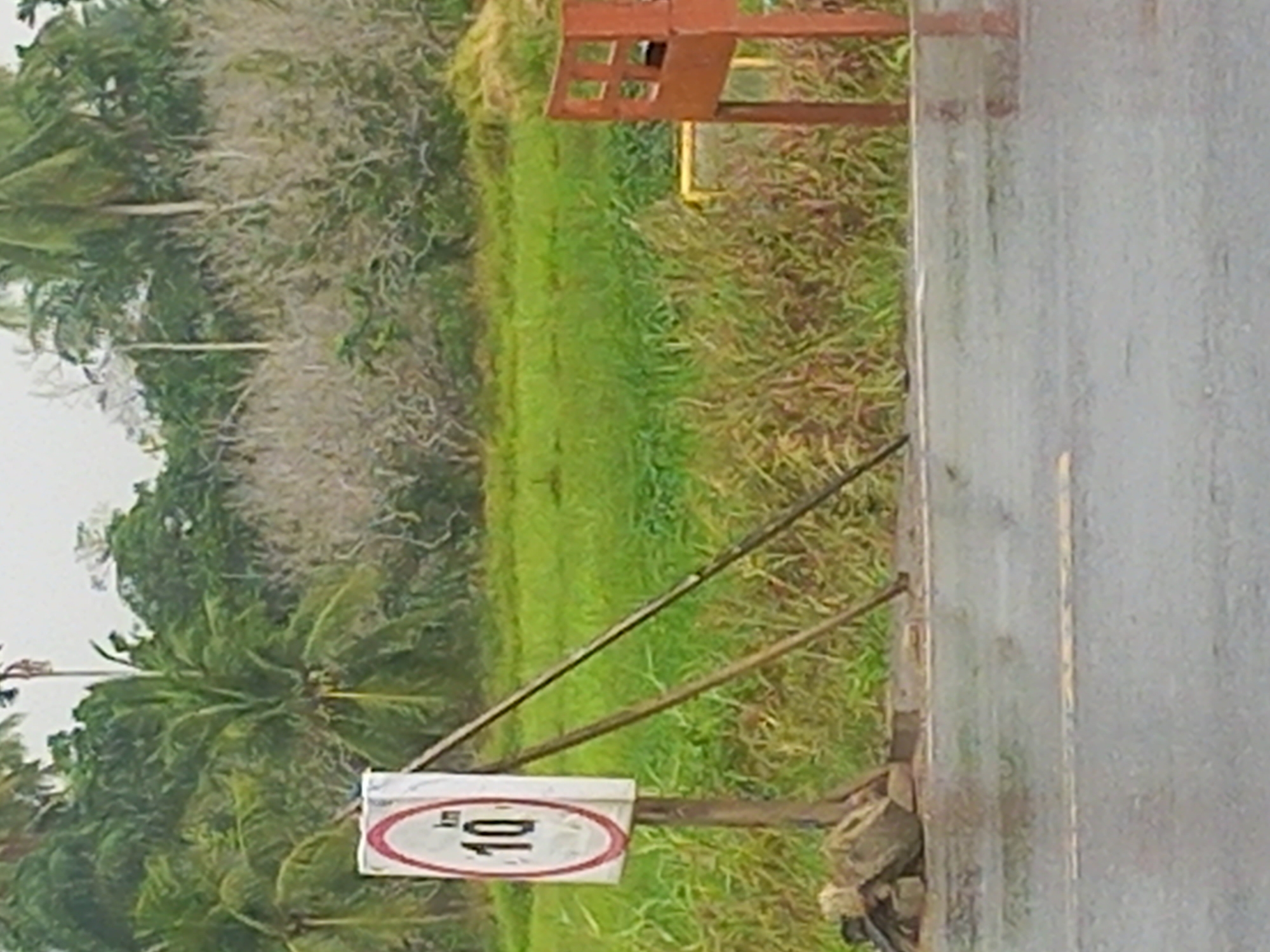 This image depicts a roadside scene in Putoh Sa, Indonesia. The focal point is a speed limit sign displaying "10 km". The sign is mounted on a wooden post, which is supported by two angled wooden planks. A pile of rocks and a damaged brick are visible at the base of the sign post. The road surface is wet, indicating it may have been raining. Beyond the road, there is a field of green grass and vegetation, with a line of trees, including palm trees, in the background. To the right of the sign, a rusty metal structure with square openings is partially visible. The sky appears overcast.