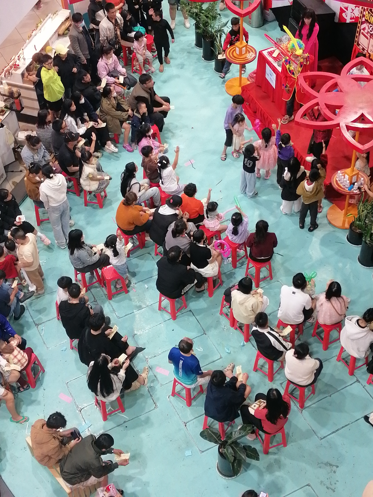 The media file captures a lively scene with a diverse group of people, predominantly children and adults, gathered in an indoor setting. The scene appears to be a public event or gathering, possibly a children's party or a promotional event, given the presence of a clown-like figure dressed in a red outfit, holding balloons and swords. Several children are excitedly watching the clown, with some raising their hands.
The setting is a brightly lit indoor space with a light blue floor, marked with white lines. Red stools are scattered throughout the area, occupied by many of the attendees who are seated, observing the proceedings. Various objects are visible, including decorative red floral structures, plants in pots, and what appears to be a small stage or performance area with a red backdrop. Some individuals are holding papers or tickets, suggesting participation in an activity or a prize draw.
The overall mood seems to be one of engagement and enjoyment, with many people focused on the central activity. The presence of balloons, colorful decorations, and the energetic performance of the clown contribute to a festive atmosphere. The time of day is not explicitly indicated, but the indoor lighting suggests it could be daytime or early evening. No specific weather conditions are visible.
While there's no discernible text in the main image that is clearly legible, the context of Da Lat, Vietnam, implies a Southeast Asian setting. The attire of the people, such as t-shirts, casual wear, and some traditional clothing elements, is consistent with the region. The image effectively conveys a sense of community gathering and shared entertainment.