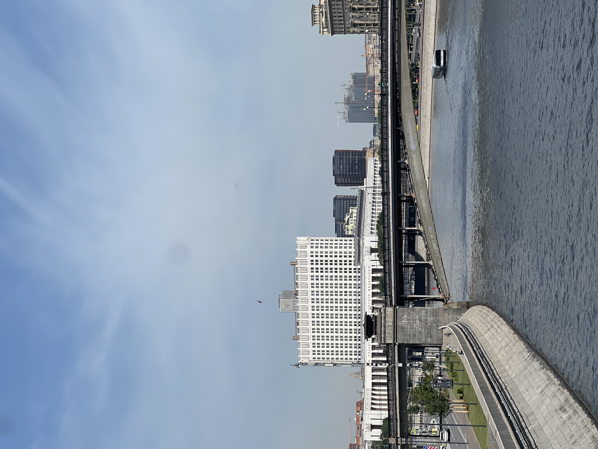 This image captures a panoramic view of the Moskva River and its surrounding urban landscape in Moscow, Russia, prominently featuring the Government House of the Russian Federation (Russian White House).
The scene is set under a clear, bright blue sky with sparse, wispy white clouds. In the foreground, the dark blue-grey waters of the Moskva River flow from left to right, exhibiting ripples and reflections of the sky. A modern, sleek white and dark grey river tram navigates the river towards the right side of the frame.
Along the left bank, a textured, light grey concrete embankment forms a curved wall, separating the river from a paved walkway and a strip of manicured green lawn dotted with small trees and streetlights. A single white car is visible on the road running parallel to the river. Several pedestrians are sparsely distributed along the embankment walkway.
Dominating the midground is a multi-level bridge structure with substantial grey stone pillars supporting upper and lower deck spans. Above and behind this bridge, the iconic white facade of the Government House of the Russian Federation stands prominently. This large, multi-story building features numerous rectangular windows arranged in a grid pattern. Its facade bears an inscription, partially visible as "ДОМ ПРАВИТЕЛЬСТВА РОССИЙСКОЙ ФЕДЕРАЦИИ" (House of the Government of the Russian Federation), and a Russian Federation flag flies atop a central tower.
To the right of the White House, a cluster of dark, modern high-rise buildings with reflective surfaces is visible. Further in the background, several tall construction cranes indicate ongoing development. To the far right, an older, ornate building with classical architectural elements completes the skyline. The overall lighting suggests a bright, sunny day.