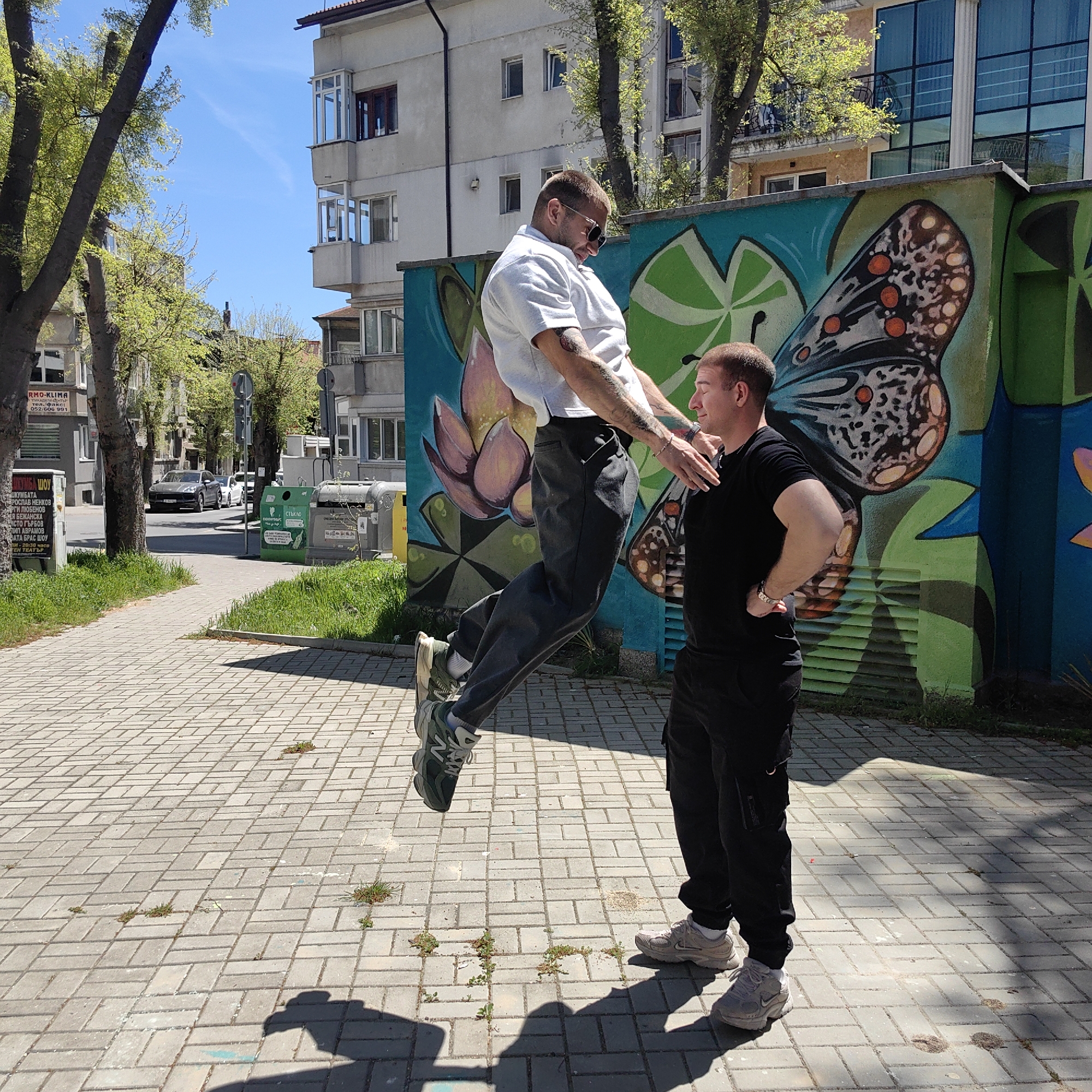 The image captures two men engaged in an outdoor activity on a sunny day in an urban setting, likely Varna, Bulgaria. One man, wearing a white short-sleeved polo shirt, dark trousers, and sunglasses, is suspended in mid-air, seemingly mid-jump or performing an acrobatic move. He has tattoos on his forearms and wears green sneakers. Facing him, the second man, dressed in a black t-shirt, black cargo pants, and light grey sneakers, stands firmly on the paved sidewalk with his hands on his hips, appearing to be observing or participating in the activity. Both men seem focused and relaxed.
The background features multi-story residential buildings with balconies, indicative of Eastern European architecture. Lush green trees line the street, suggesting spring or early summer. Immediately behind the men is a vibrant wall covered in colorful graffiti depicting a large butterfly and floral patterns in blues, greens, and oranges. The scene includes a paved sidewalk with patches of grass, and in the distance, a street with parked cars. Visible objects include green and yellow recycling bins labeled with Cyrillic text, specifically "стъкло" (glass), and an advertising board also bearing Cyrillic text promoting a "TEAT" (theater) event or show with names like "КРОСЛАВ НЕНКОВ" and "АВРАМОВ". The clear blue sky and distinct shadows cast on the ground indicate bright daylight, likely midday or early afternoon.