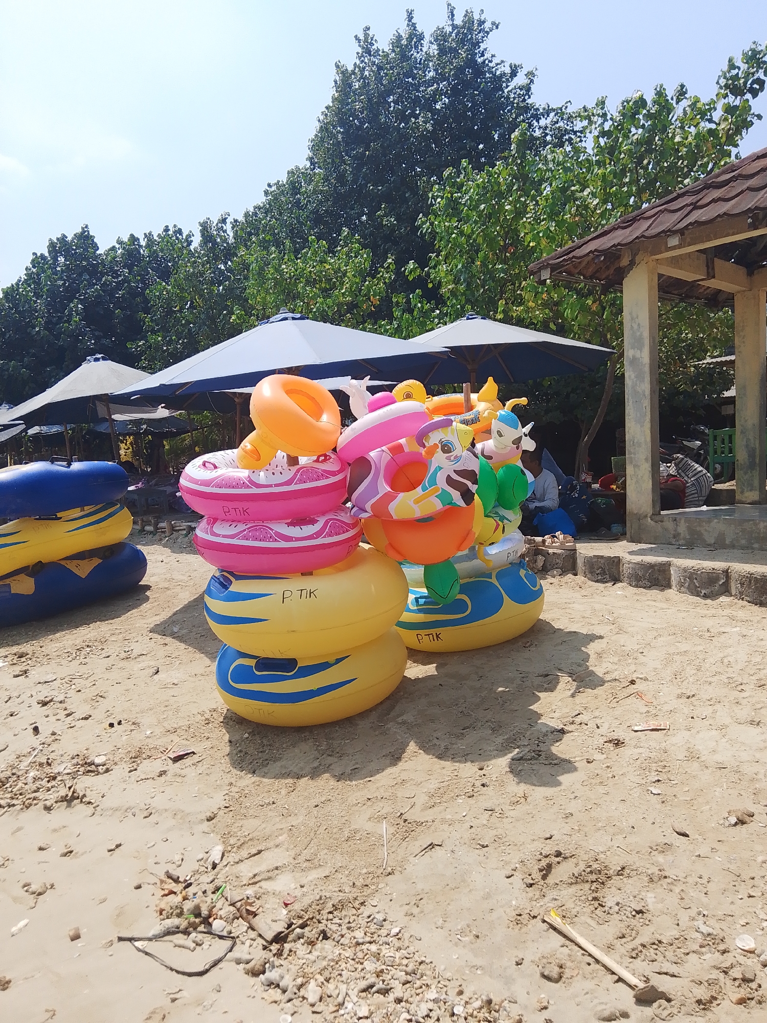 The image depicts a beach scene in Tegalsambi, Indonesia, on a clear, sunny day. In the foreground, a collection of colorful inflatable swimming rings and pool floats are stacked on the sand. Some of the inflatables are circular rings with patterns, while others are shaped like cartoon characters. To the left of the inflatables, more blue and yellow inflatable rafts are stacked. In the background, several large blue umbrellas are set up on the sand, providing shade. Behind the umbrellas, lush green trees form a dense backdrop against the sky. To the right, a simple, open-air structure with a tiled roof stands on a raised platform. Underneath this structure, a few individuals are seated or resting, with some bags and motorcycles visible. The ground is covered in sand and small shells, with scattered debris like dried leaves and small plastic items.