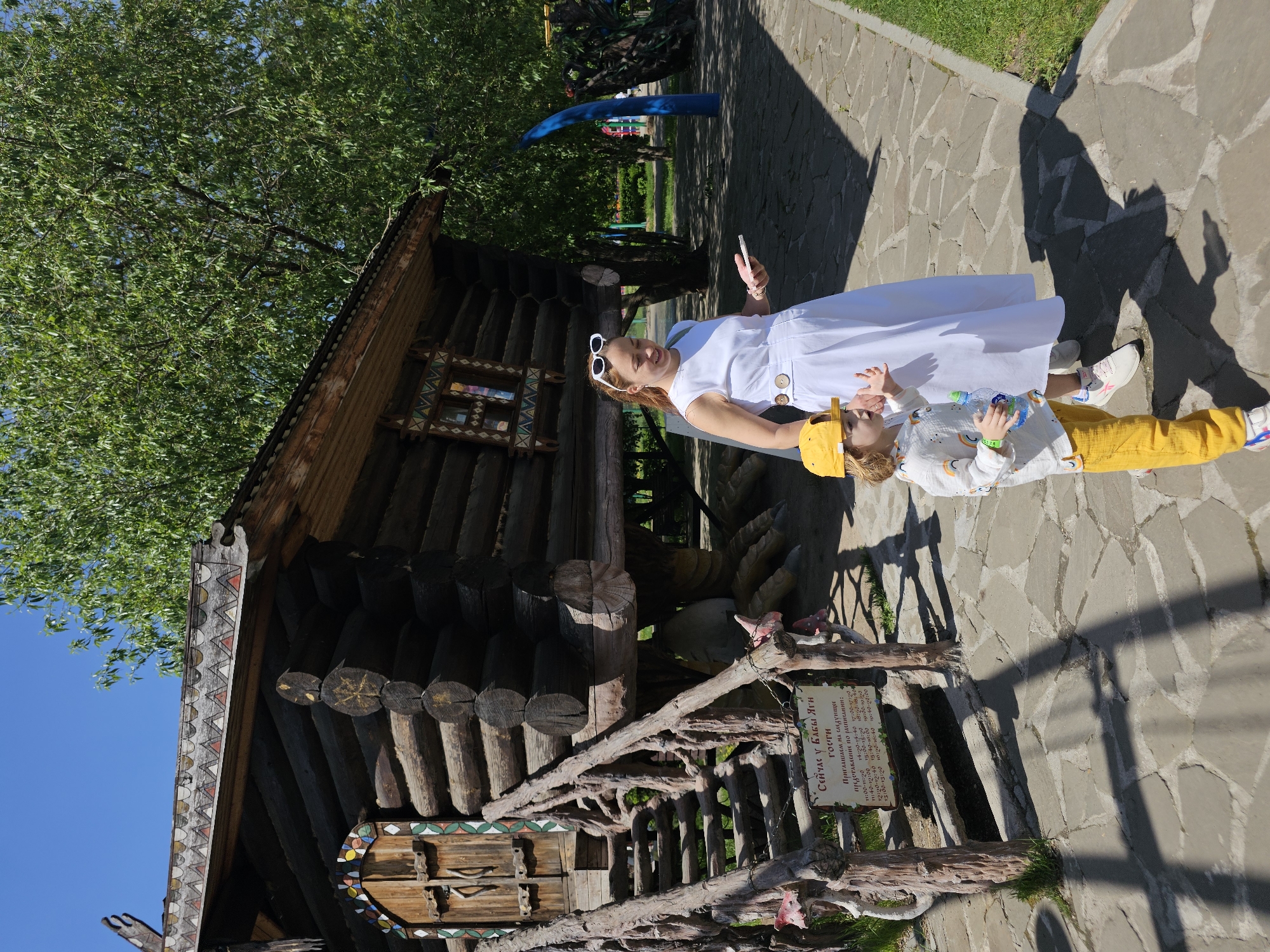 Here's a detailed description of the image:
**Scene:** A woman and child are standing outside a wooden structure that resembles a Russian fairytale house, likely the Baba Yaga's house. The woman, wearing a white dress and sunglasses, holds a phone. The child, wearing yellow pants and a yellow hat, holds a water bottle.
**Environment:** The scene takes place on a stone-paved path with grass visible at the edges. The day is sunny, casting shadows. Trees and greenery are visible in the background. A small sign is posted nearby.
**People:**
* A woman in a white dress and sunglasses
* A child in yellow pants and a yellow hat
**Objects:**
* Baba Yaga's wooden house
* Stone path
* Trees
* Greenery
* Phone
* Water bottle
* Sign with Cyrillic text
**Interactions:** The woman is holding a phone and appears to be taking a picture or video. The child is holding a water bottle.
**Location:** The Cyrillic text on the sign mentions Baba Yaga, suggesting the scene is likely a themed area related to Russian folklore. The specific location is Khimki, Russia, as provided.