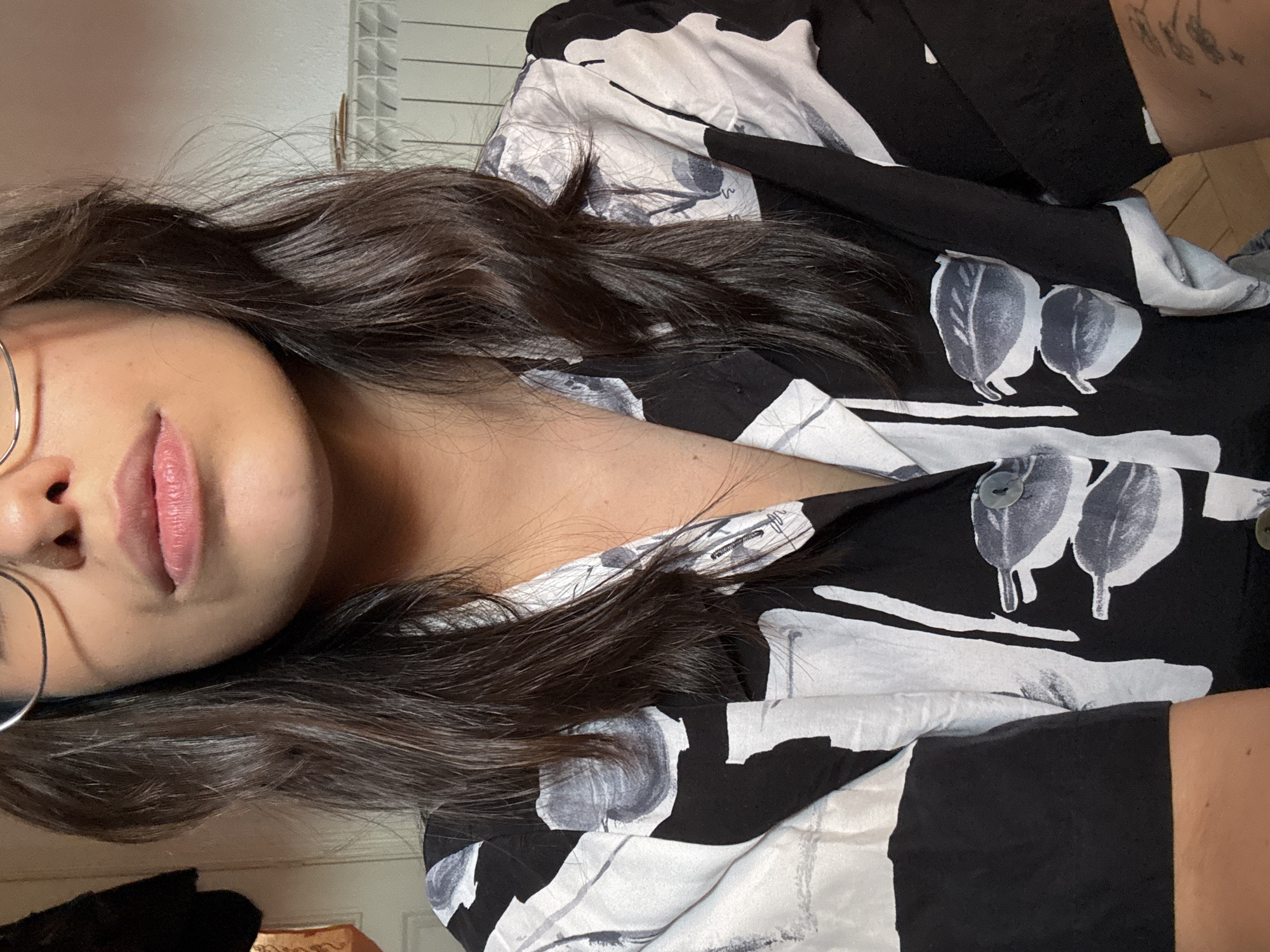 A close-up, front-facing shot focuses on the upper torso and face of a person wearing glasses and a black and white patterned shirt. The shirt has a collared neckline and a button-up front with a visible button. The person's dark, shoulder-length hair frames their face, and their lips are slightly parted. The background is a plain, light-colored wall with a white radiator visible on the right side. The image does not contain any information to determine the location in the city of Las Rozas de Madrid, Spain.