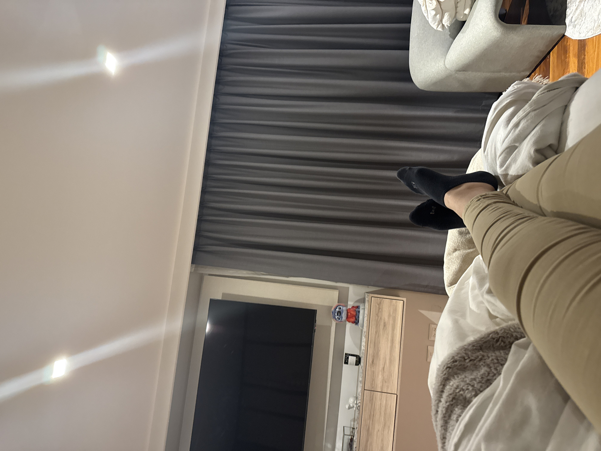 A person wearing tan pants and black socks is relaxing in bed in a room with gray curtains. A gray chair is visible in the background. A television and a wooden cabinet are near the bed. The room appears to be well-lit with overhead lighting. No evidence suggests this photo was taken in San Bernardino Tlaxcalancingo, Mexico.