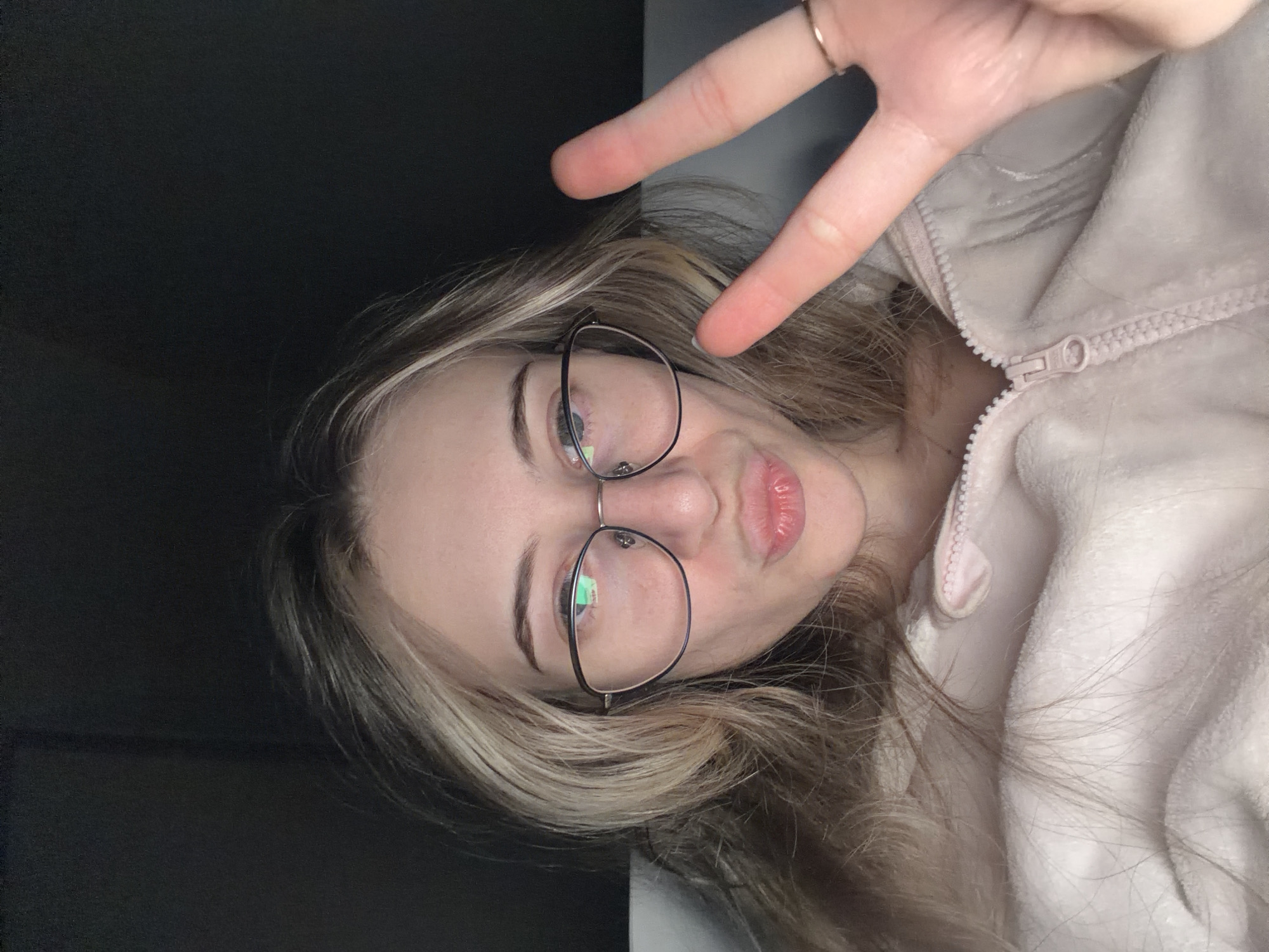 A young woman with blonde and brown hair is wearing glasses and a pink zip-up jacket. She is making a peace sign with her right hand. The background is a dark gray color. There is no evidence to suggest that this image was taken in Plovdiv, Bulgaria.