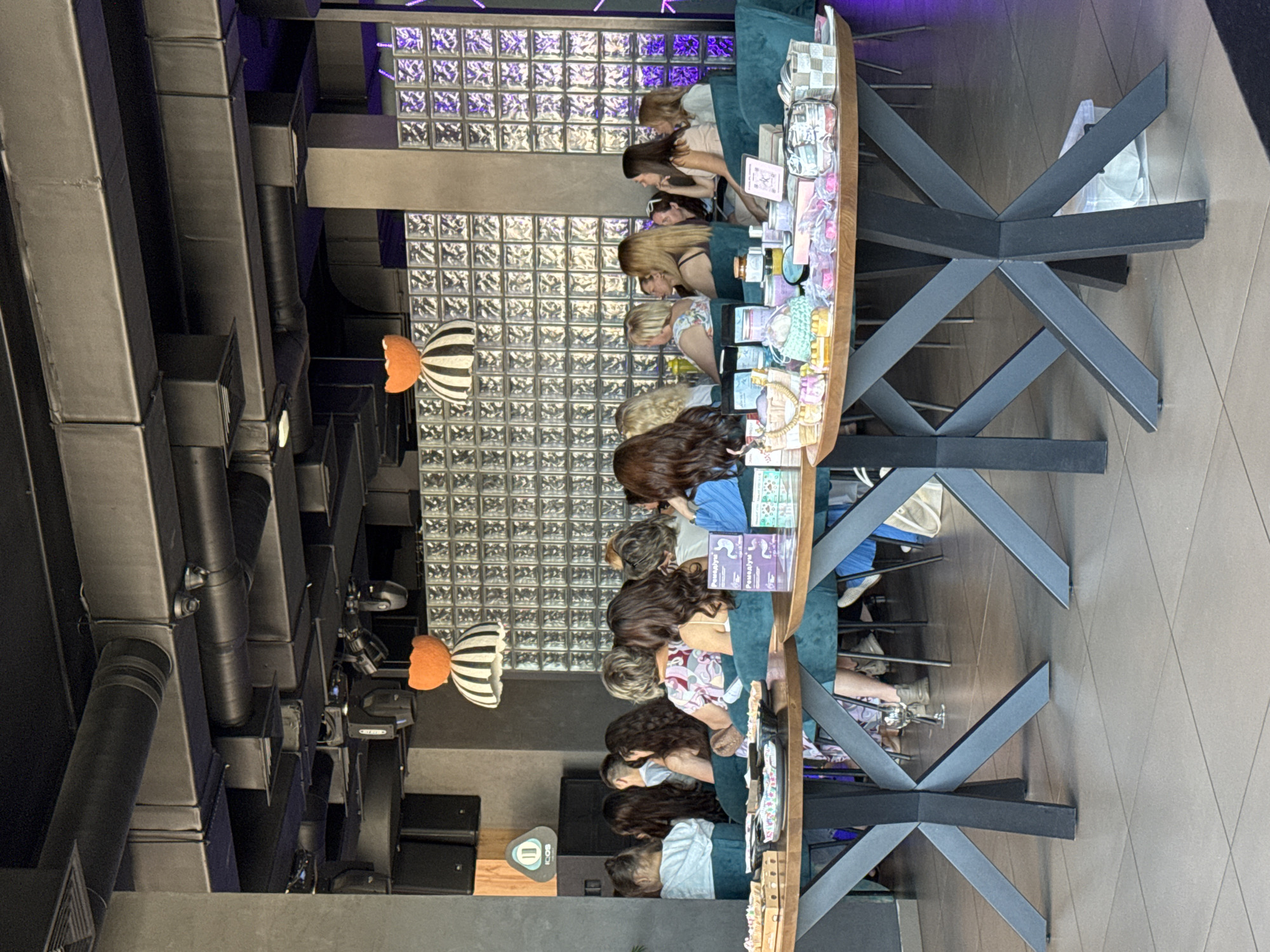 The image shows a group of women seated in a room that appears to be a venue in Kryvyi Rih, Ukraine, possibly for an event or workshop. They are seated at tables laden with various items, including what appear to be cosmetic products, gift bags, and decorative elements. The room has a modern industrial design, with exposed ductwork and lighting fixtures on the ceiling. The background features a wall of textured glass blocks with purple lighting. The women are mostly facing towards the front, suggesting they are listening to a speaker or participating in an activity. The overall atmosphere seems to be one of a social gathering or a product presentation.