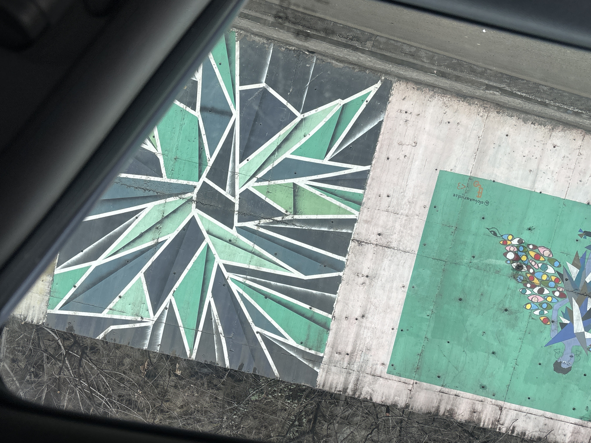 A concrete retaining wall along the right embankment of the Mtkvari River (Kura River) in Tbilisi, Georgia, specifically within or between the Saburtalo and Didube districts, is depicted from an elevated interior vehicle perspective.
The wall features two distinct murals. On the left, a rectangular green panel displays a stylized depiction of a light-blue-skinned female figure with short dark hair and triangular earrings, adorned in a multi-faceted geometric blue and white garment. A cascade of numerous multi-colored eyes, along with small dark bird-like figures, flows from her lower left side downwards and to the right. The artist's handle, "@elobakanidze," followed by a small orange key icon and a pink heart icon, is inscribed in the upper right of this green panel.
To the right, an abstract geometric mural covers a larger section of the wall. This design consists of interlocking polygons and sharp angles rendered in shades of dark gray, black, and various greens, all outlined by stark white lines, creating a radiating or fractured pattern.
The unpainted concrete sections of the wall between and around the murals are light gray, marked by darker stains, streaks, and multiple small, dark circular depressions consistent with bolt holes. Above the wall, tangled, bare tree branches and twigs are visible against a lighter sky, suggesting a natural embankment or hillside. Below the wall, a dark gray road or path runs parallel, separated by a slightly raised, dark curb. A small white object lies on the road surface directly beneath the curb. The scene is partially framed along the bottom and right edges by the dark gray interior trim of a vehicle's window.