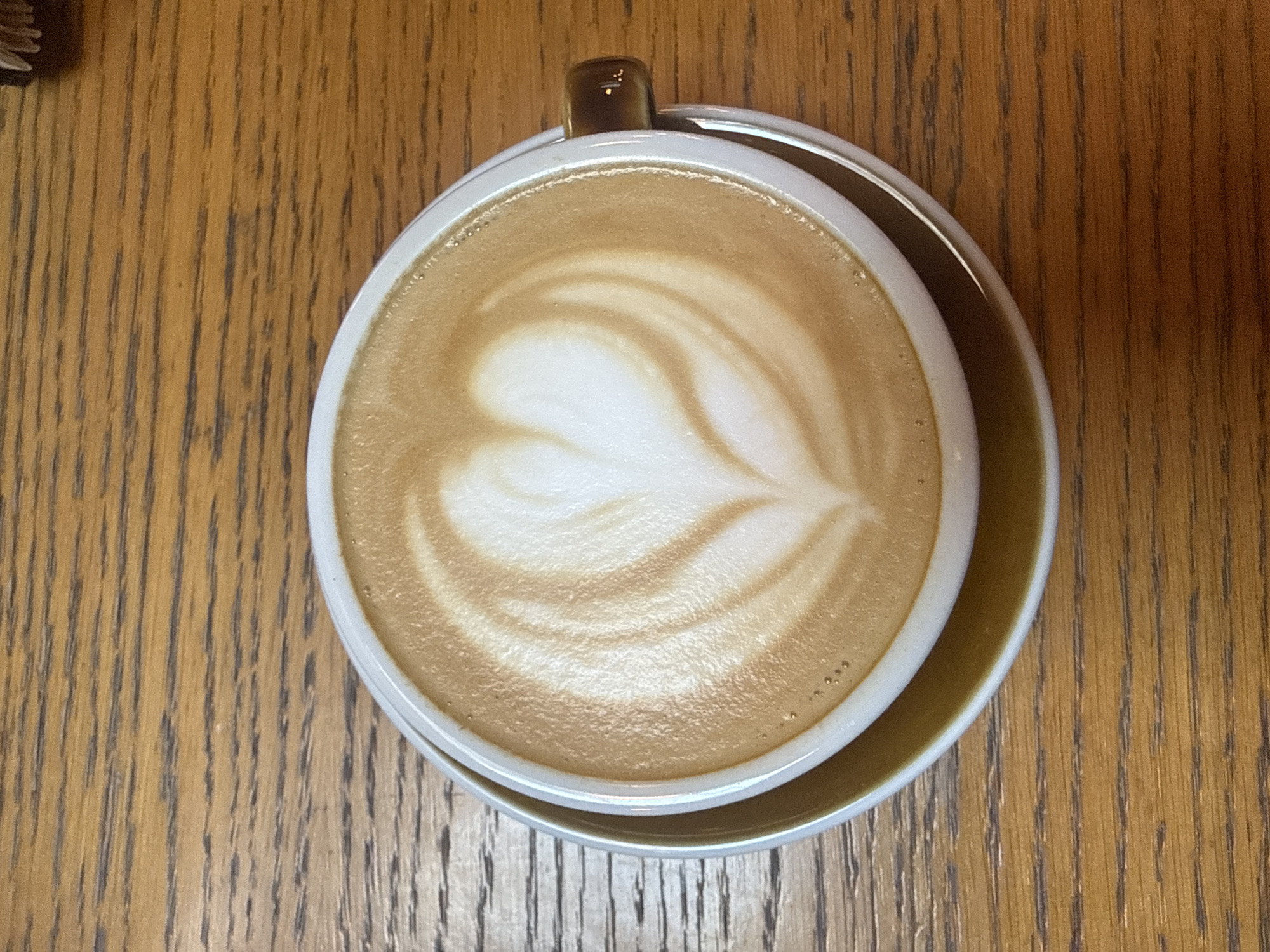 An overhead shot shows a cup of latte art in the center of an oak table. The latte art depicts a heart or tulip design. The cup is white with a brown handle and is set on a saucer with a similar color scheme to the handle. The oak table has a distinct wood grain pattern. The location is in Sofia, Bulgaria.