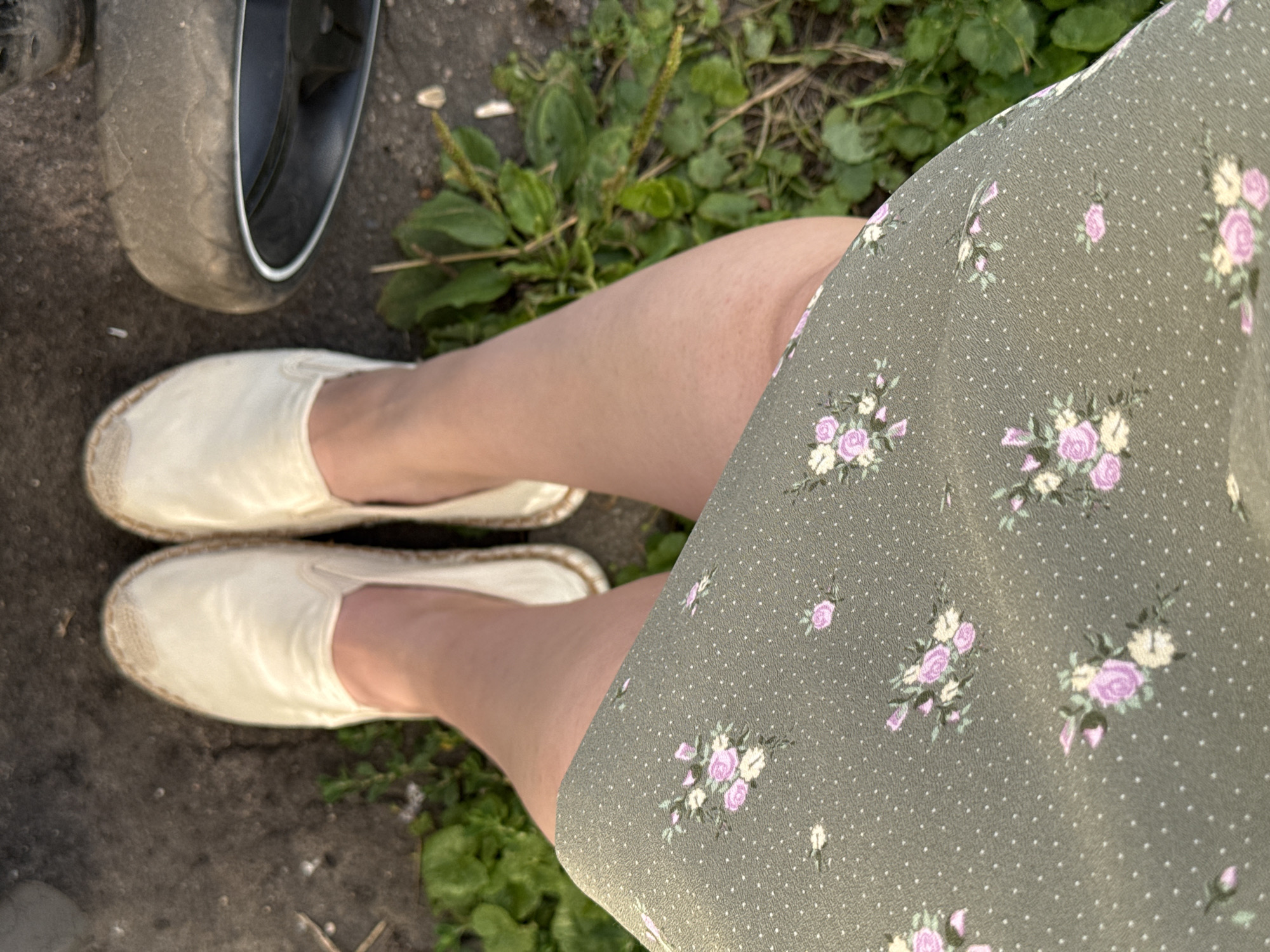 In Lyubertsy, Russia, a person stands outdoors, their legs and feet visible from the knees down. They are wearing a green floral print dress and off-white espadrille shoes. To the right, a portion of a dark-colored tire is visible, suggesting the presence of a vehicle or bicycle. The ground is a mixture of asphalt and dirt, with patches of green weeds and grass growing around the edges. The lighting indicates it is daytime, possibly late afternoon due to the warm tones.
