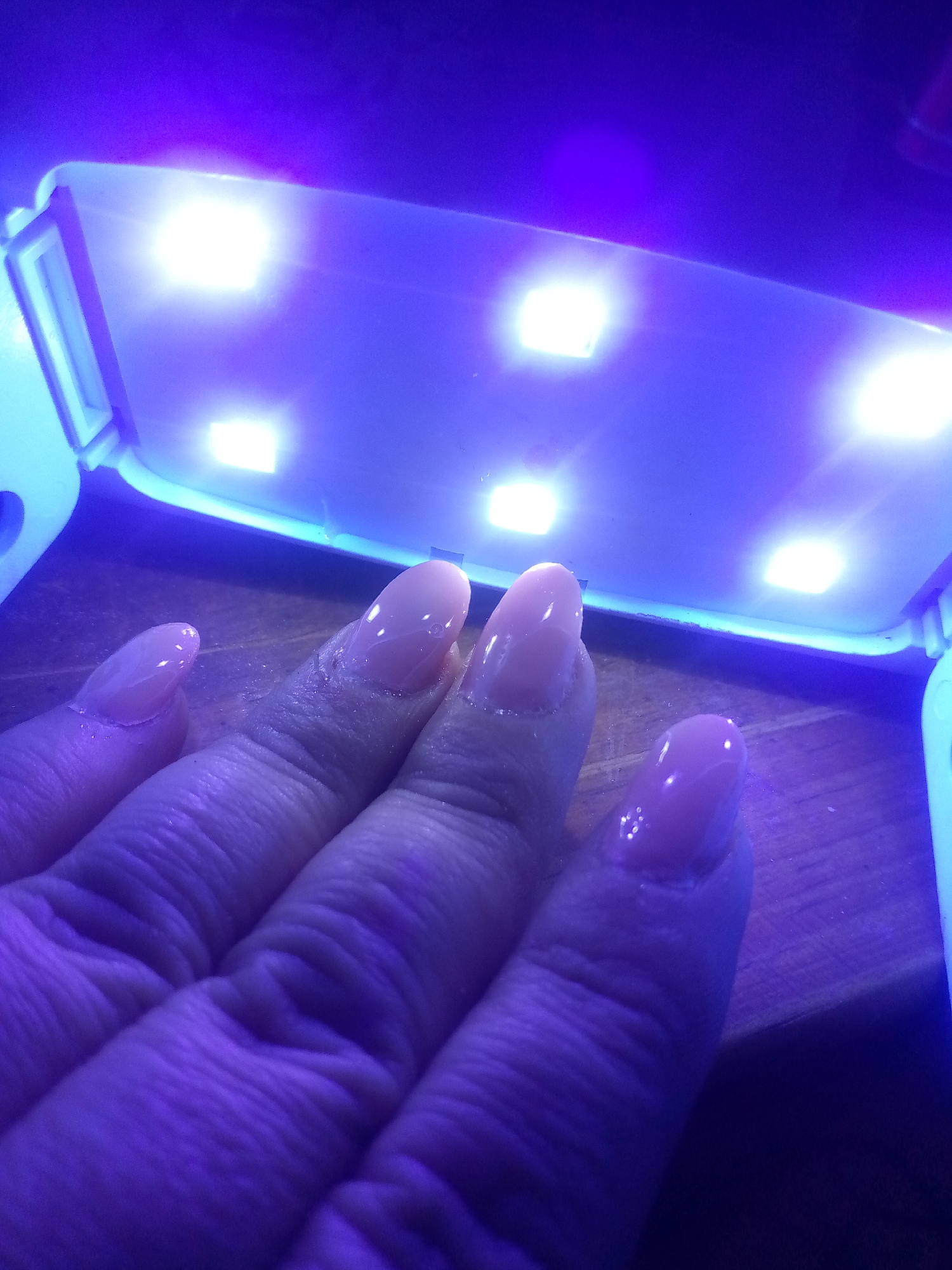 A close-up image shows a human hand with light peach colored nails under a UV nail curing lamp. The lamp has multiple bright LED lights. The hand is positioned directly underneath the lamp. The background appears to be a wooden surface. No information indicates the location of the image as Juan Griego, Venezuela.