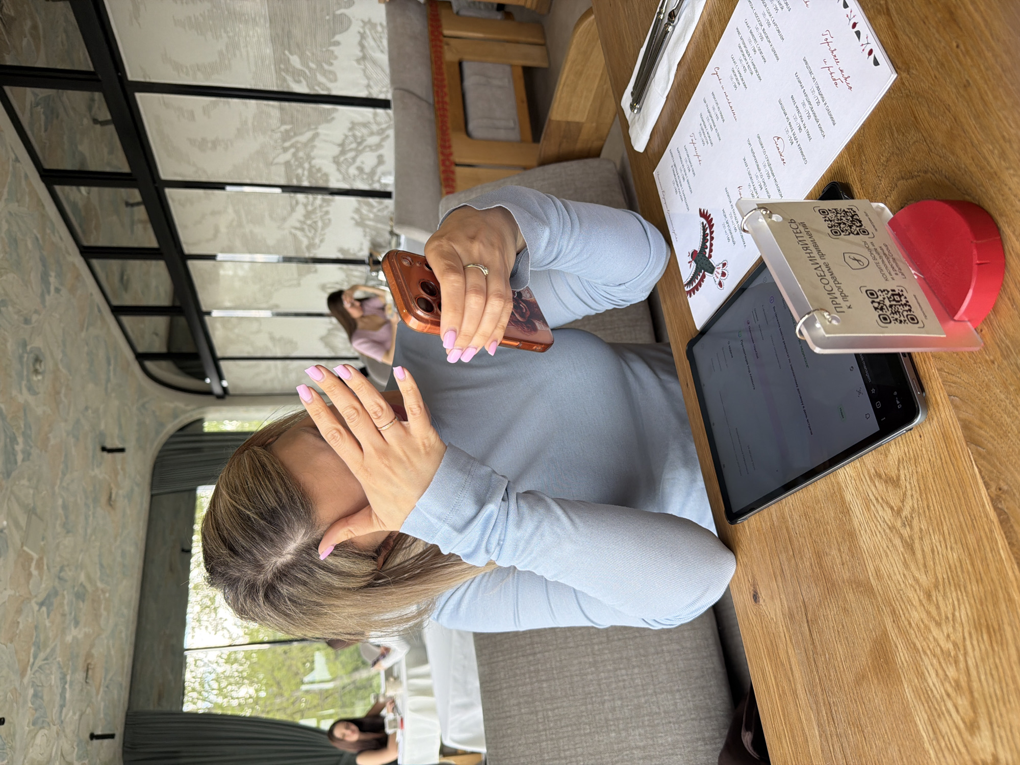 A woman with shoulder-length blonde hair, wearing a light blue long-sleeved shirt and rings on her fingers, is seated at a wooden table in a restaurant. She is holding a cell phone in her right hand and appears to be covering her face with her left hand. On the table, there is a menu with Cyrillic text, a tablet, a holder with QR codes, and cutlery. A window is visible behind the woman. Another woman is visible in the background. The restaurant has patterned wallpaper and a metal-framed ceiling. The scene is located in Krasnoyarsk, Russia.